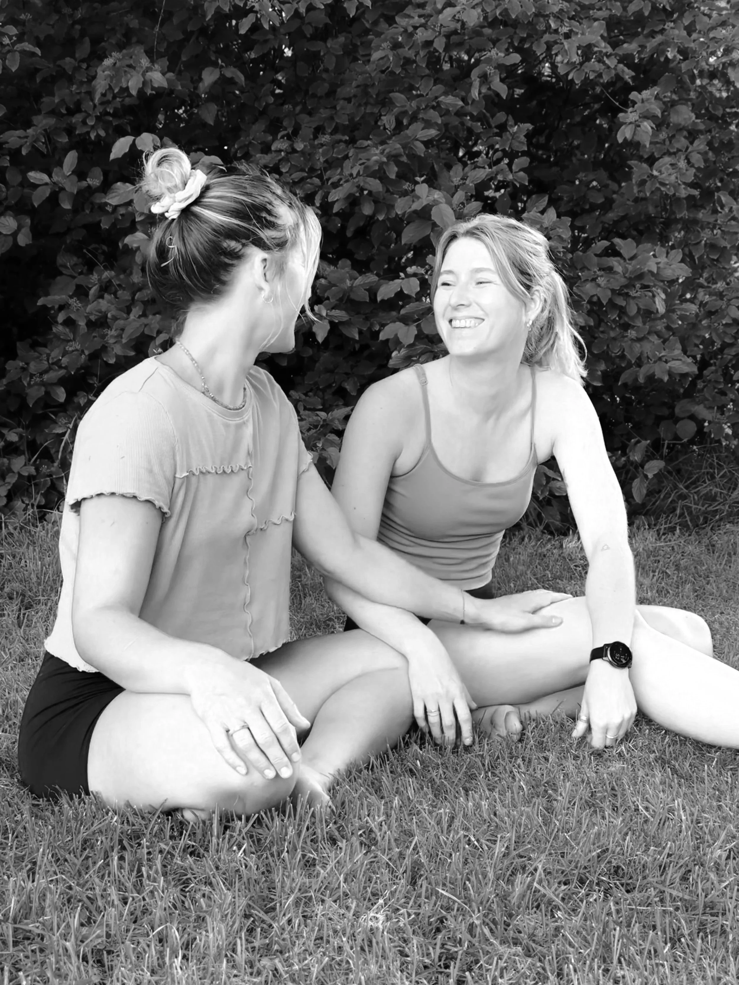 Zwei Frauen sitzen auf dem Gras und lachen miteinander, im Hintergrund Büsche und Bäume.