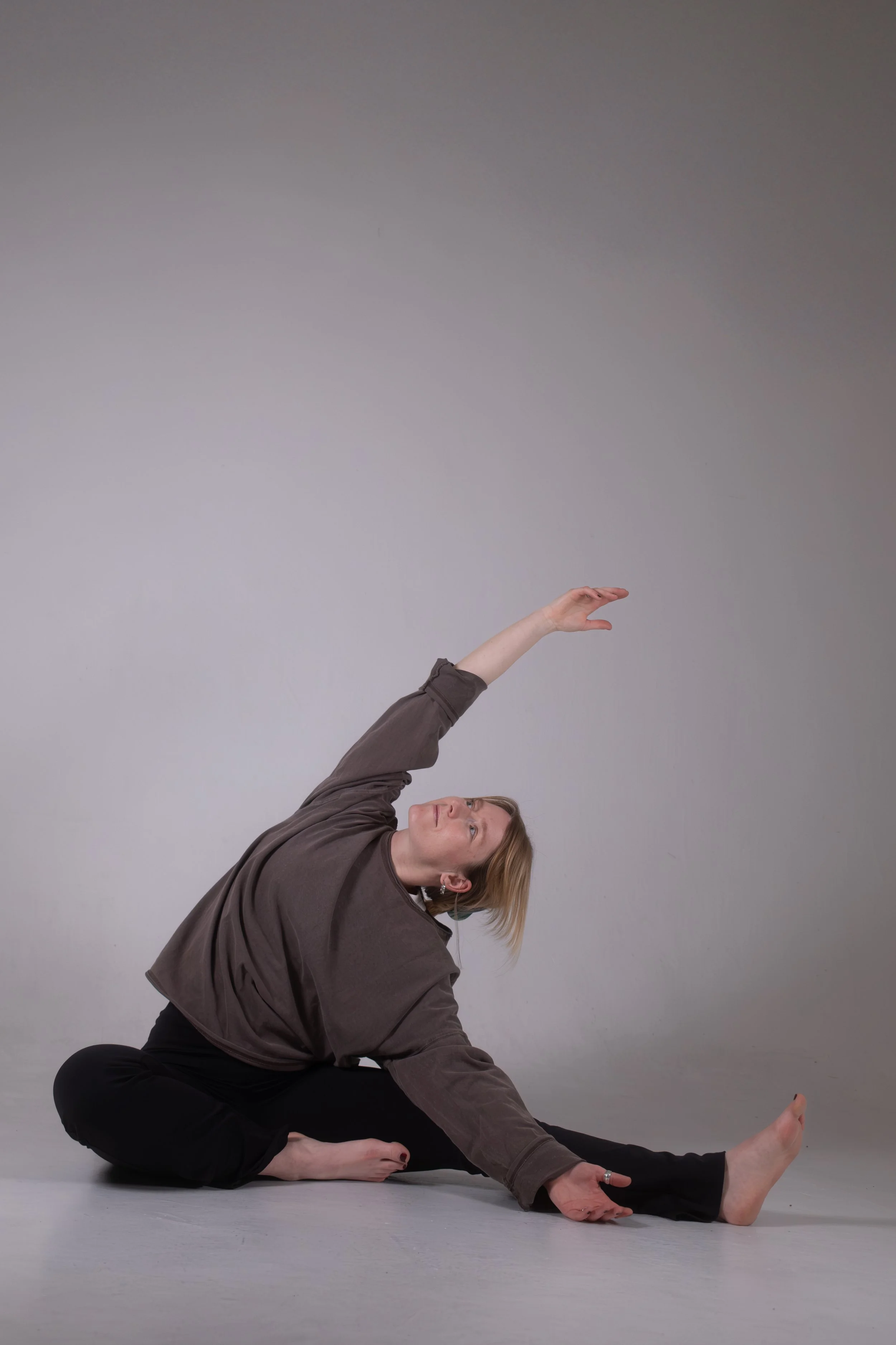 Eine Frau in schwarzen Hosen und braunem Pullover führt eine Yoga- oder Dehnübung auf dem Boden aus, während sie nach oben und zur Seite streckt, vor einem grauen Hintergrund.