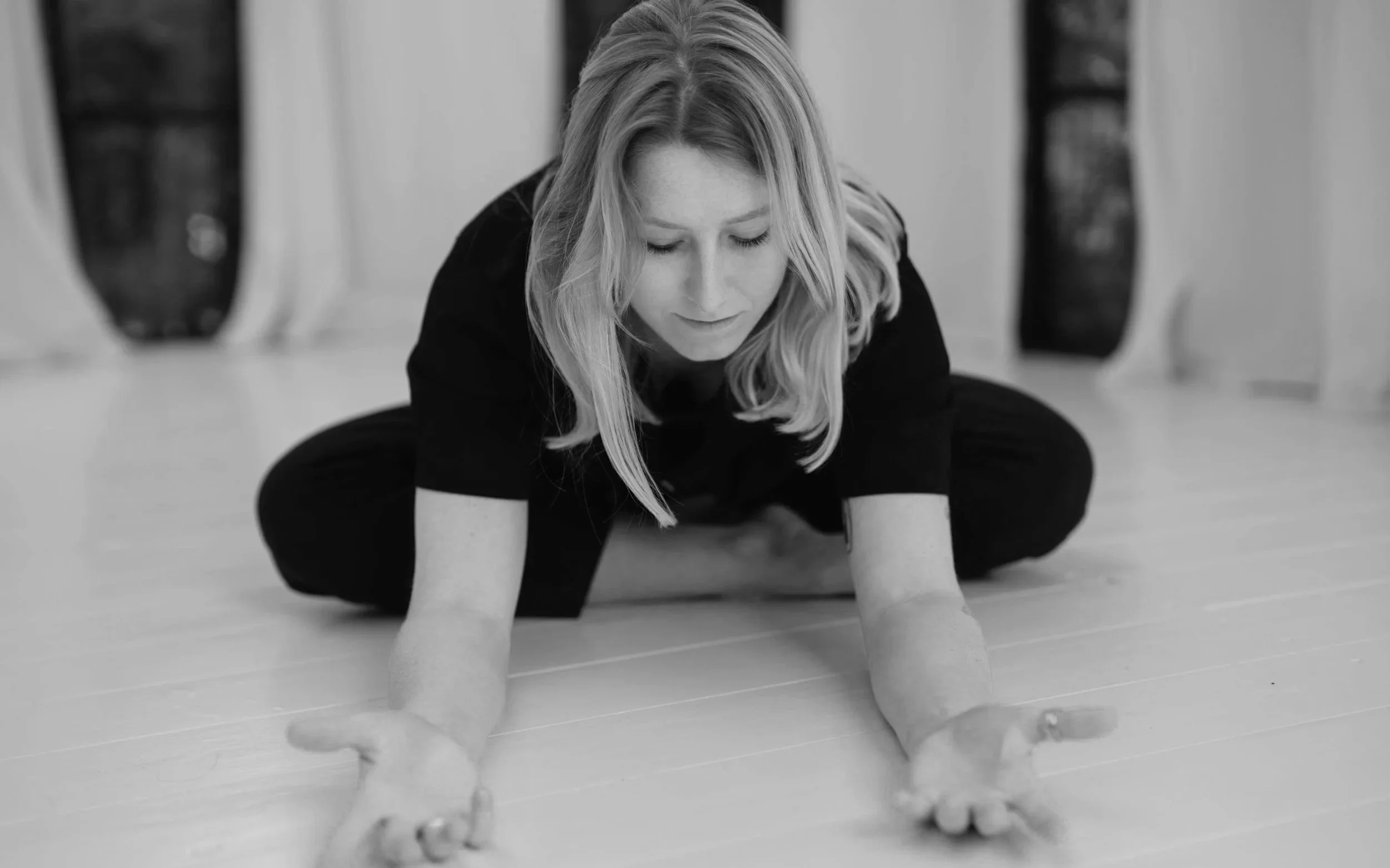 Frau macht Yoga auf dem Boden, blick nach unten, mit geschlossenen Augen, in einem hellen Raum mit großen Fenstern im Hintergrund.
