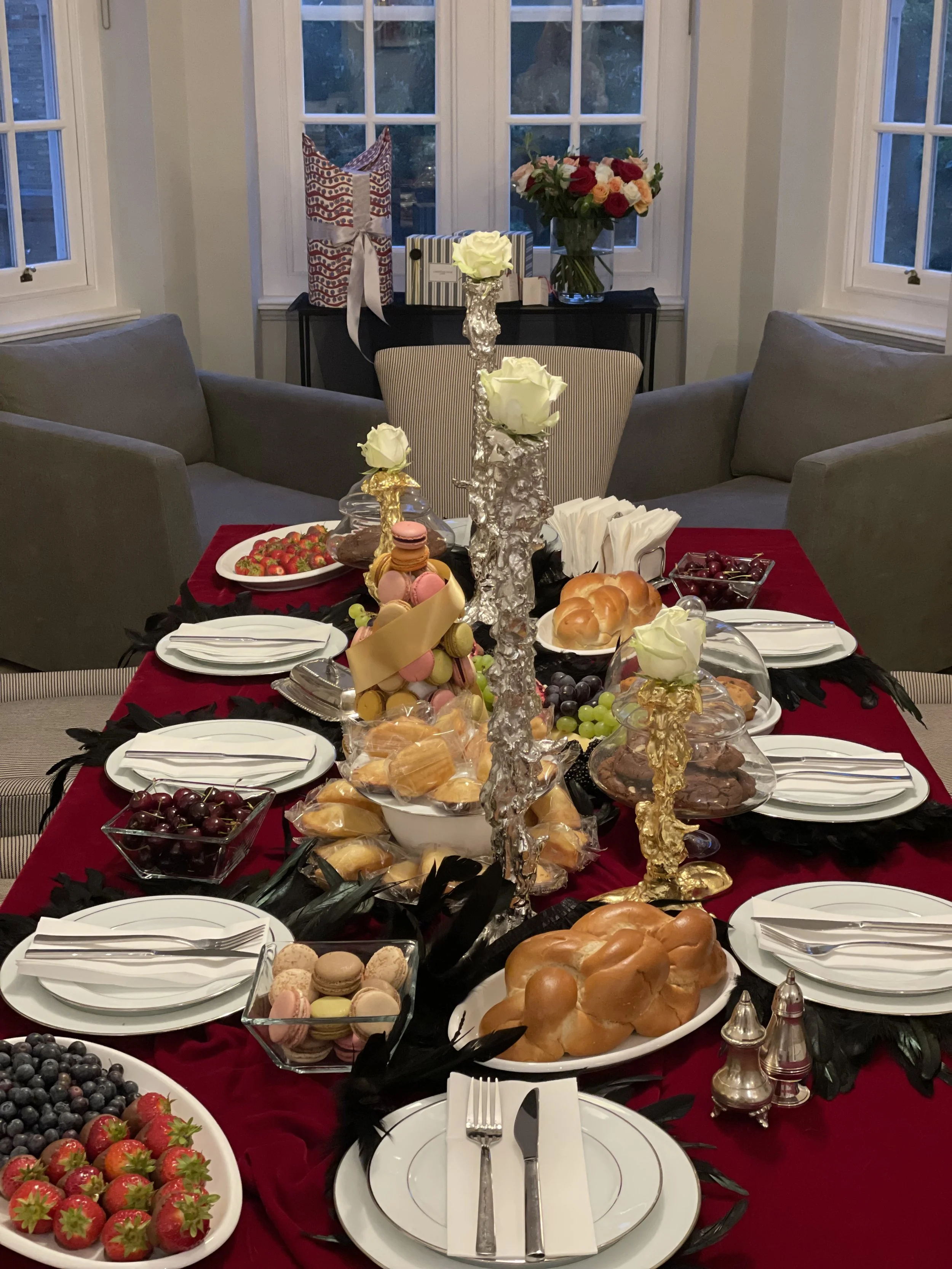 Elegant dining table set with a red tablecloth, featuring plates of fruit, pastries, macarons, and rolls, with ornate candlesticks and white roses as centerpieces.