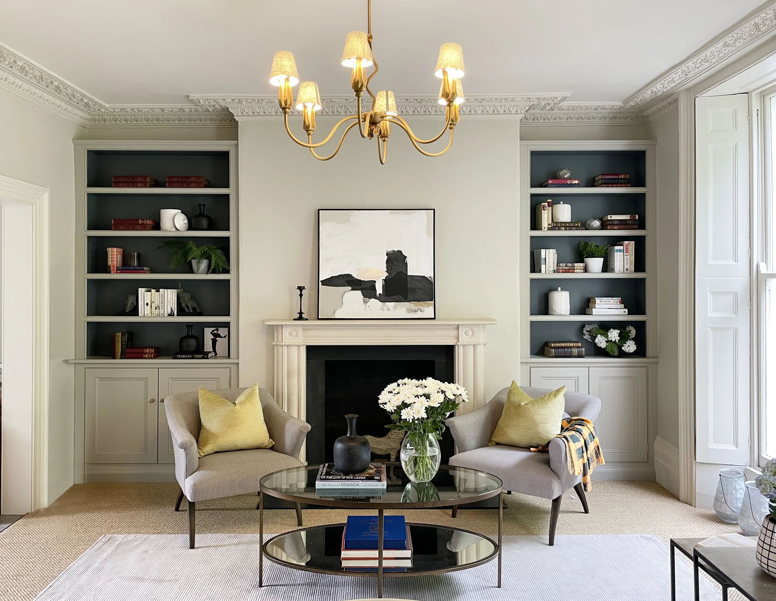 Elegant living room with neutral tones, featuring a fireplace with white mantel, abstract painting above it, two light gray armchairs with yellow pillows, glass coffee table with flowers and books, and built-in shelves filled with decorative items and books on either side of the fireplace.