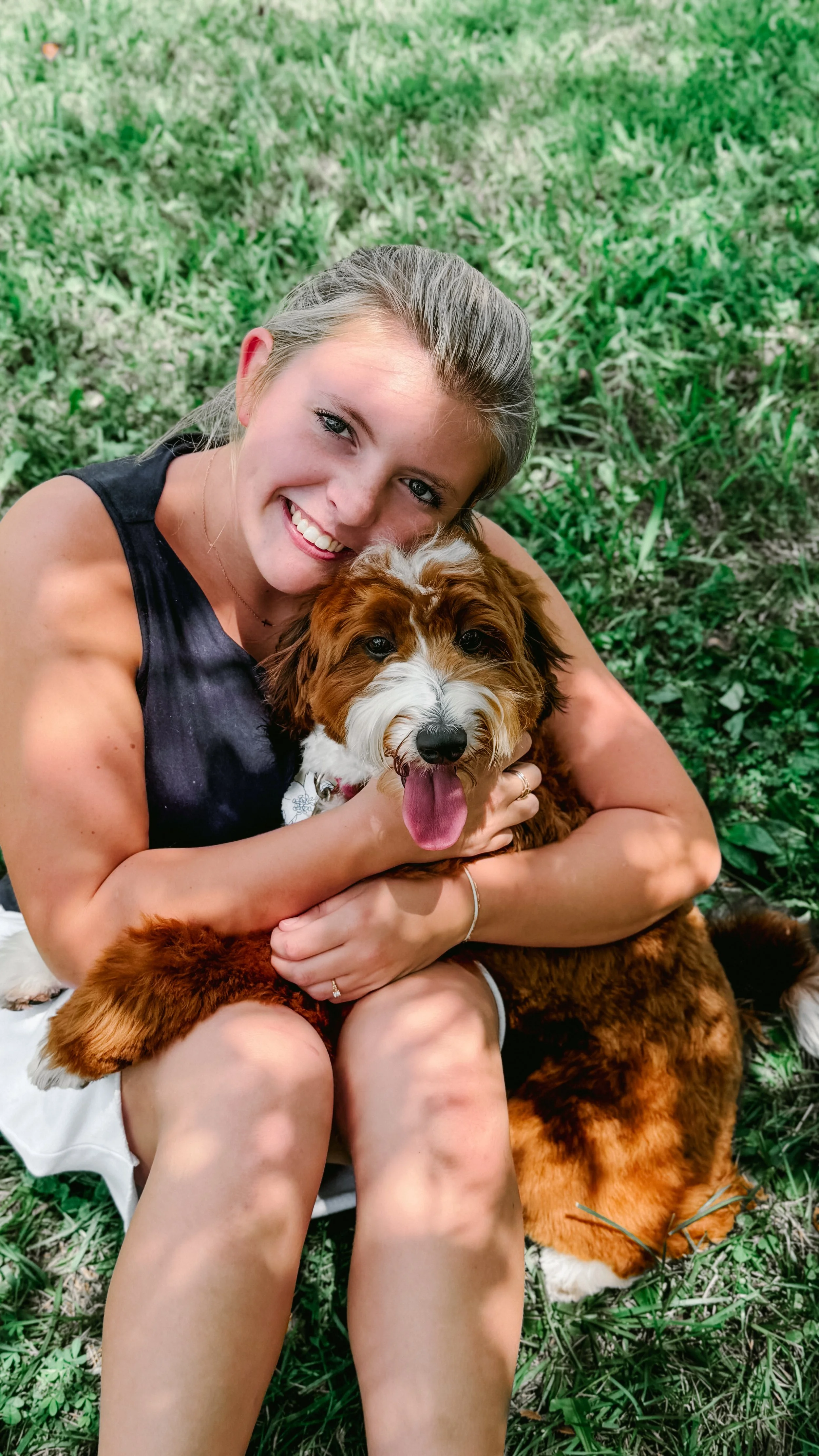 A woman with blonde hair and a black sleeveless top sitting outdoors on grass, smiling and holding a fluffy brown and white dog with its tongue out.