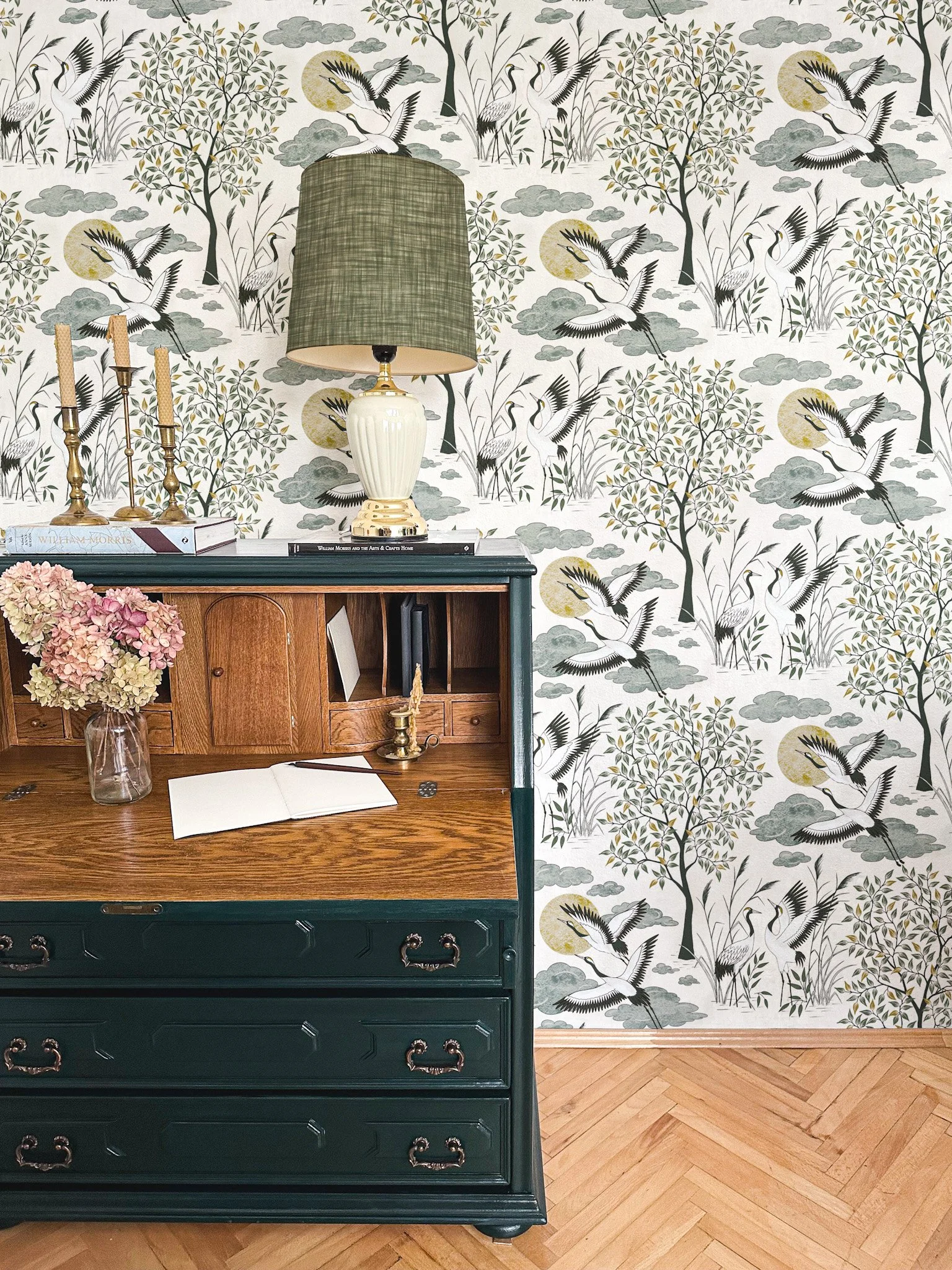 A vintage wooden desk with green drawers, decorated with a vase of pink and white hydrangeas, an open notebook, a pen, a small brass candlestick, and a brass ashtray. Behind the desk is wallpaper featuring cranes flying among trees and clouds, with a full moon in the background. A table lamp with a green shade and a white ceramic base sits atop the desk, along with books and brass candlesticks.