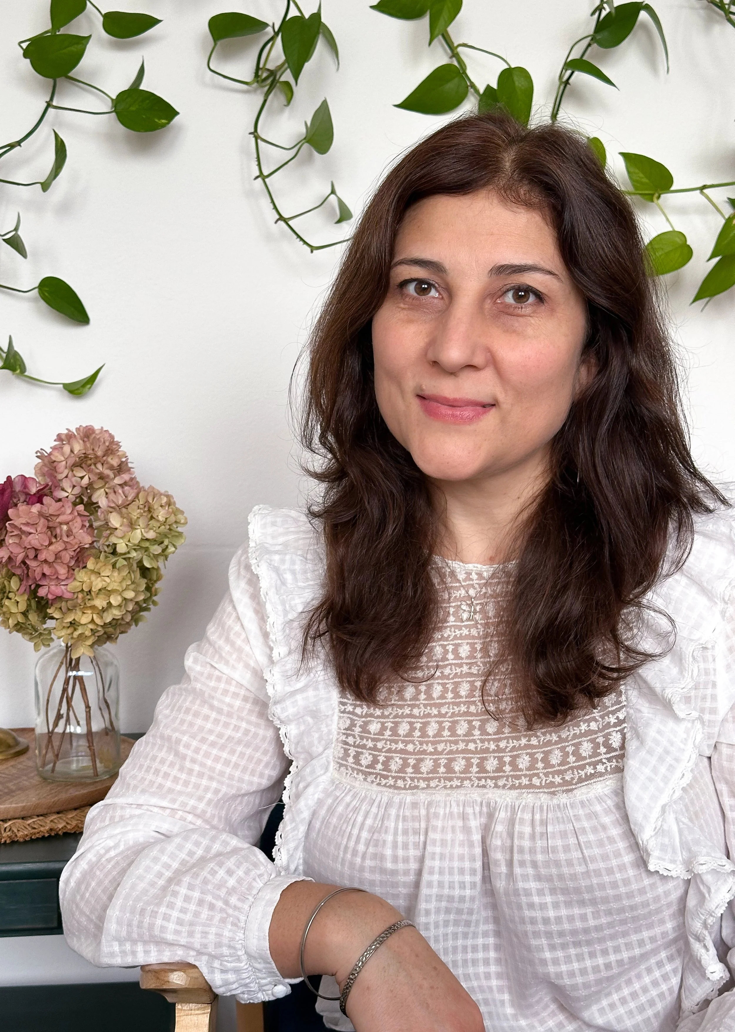 A woman with long brown wavy hair, wearing a white embroidered blouse, sitting indoors with a white wall, green leafy plants, and a vase of dried pink and green hydrangeas in the background.