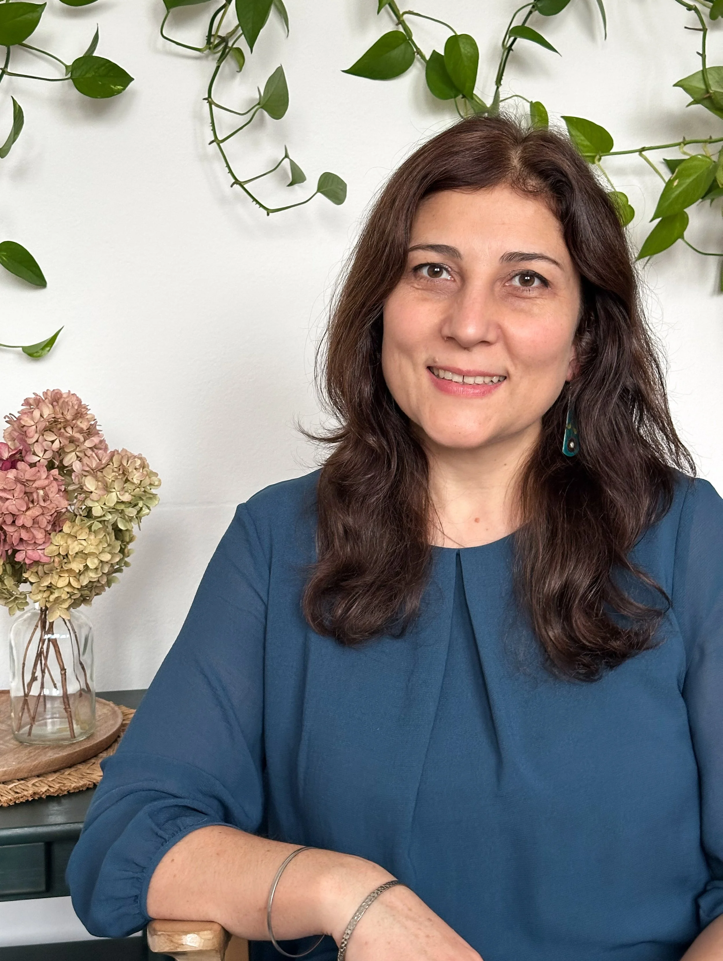 A woman with long brown hair and light skin, smiling, wearing a blue blouse, with greenery behind her and a vase of pink and beige hydrangeas on a wooden table.