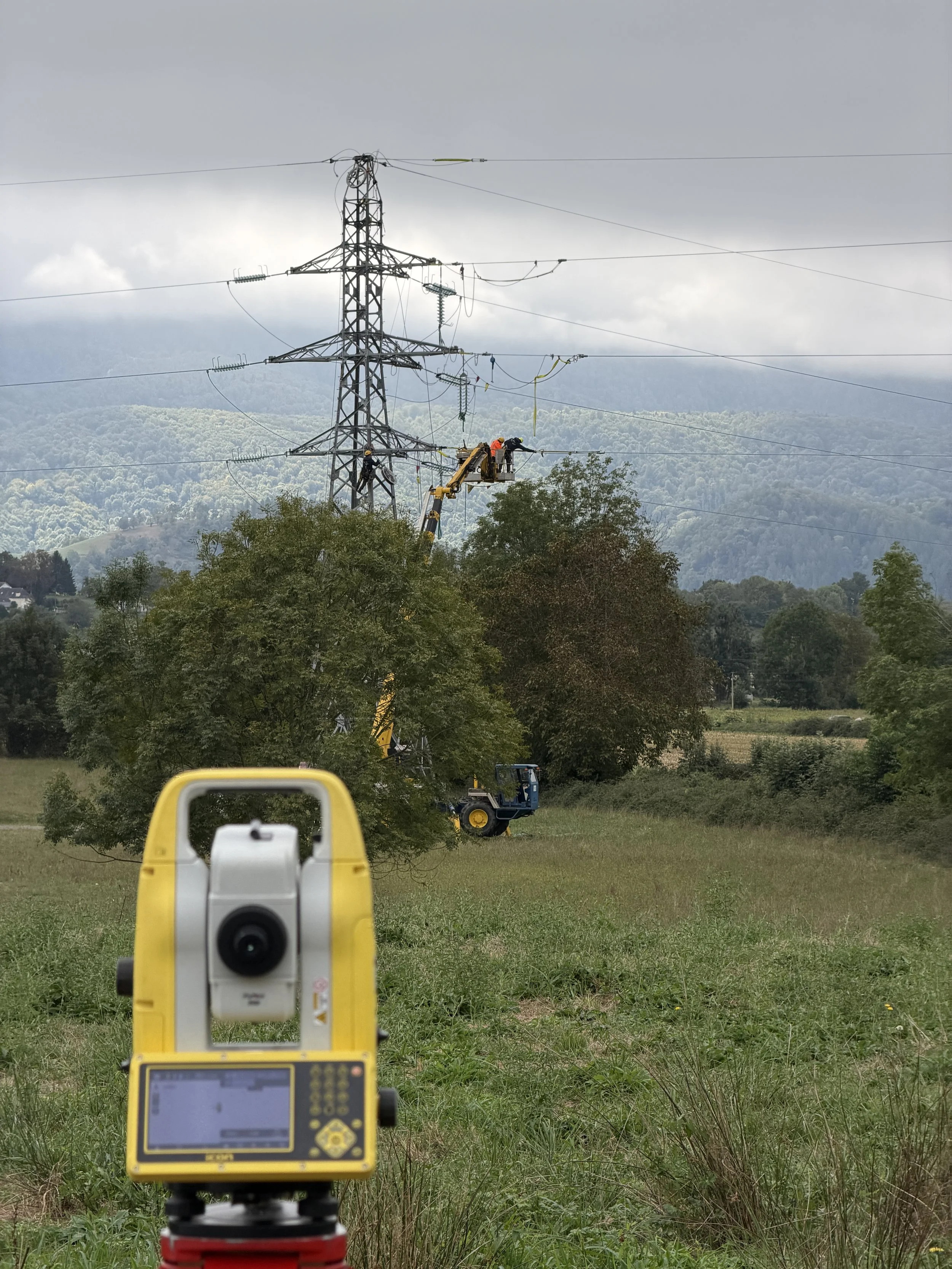 Travaux sur une ligne électrique haute tension à l'aide d'appareil topographique de qualité, au milieu d'une zone rurale avec des collines en arrière-plan, sous un ciel nuageux.
Lebon Services