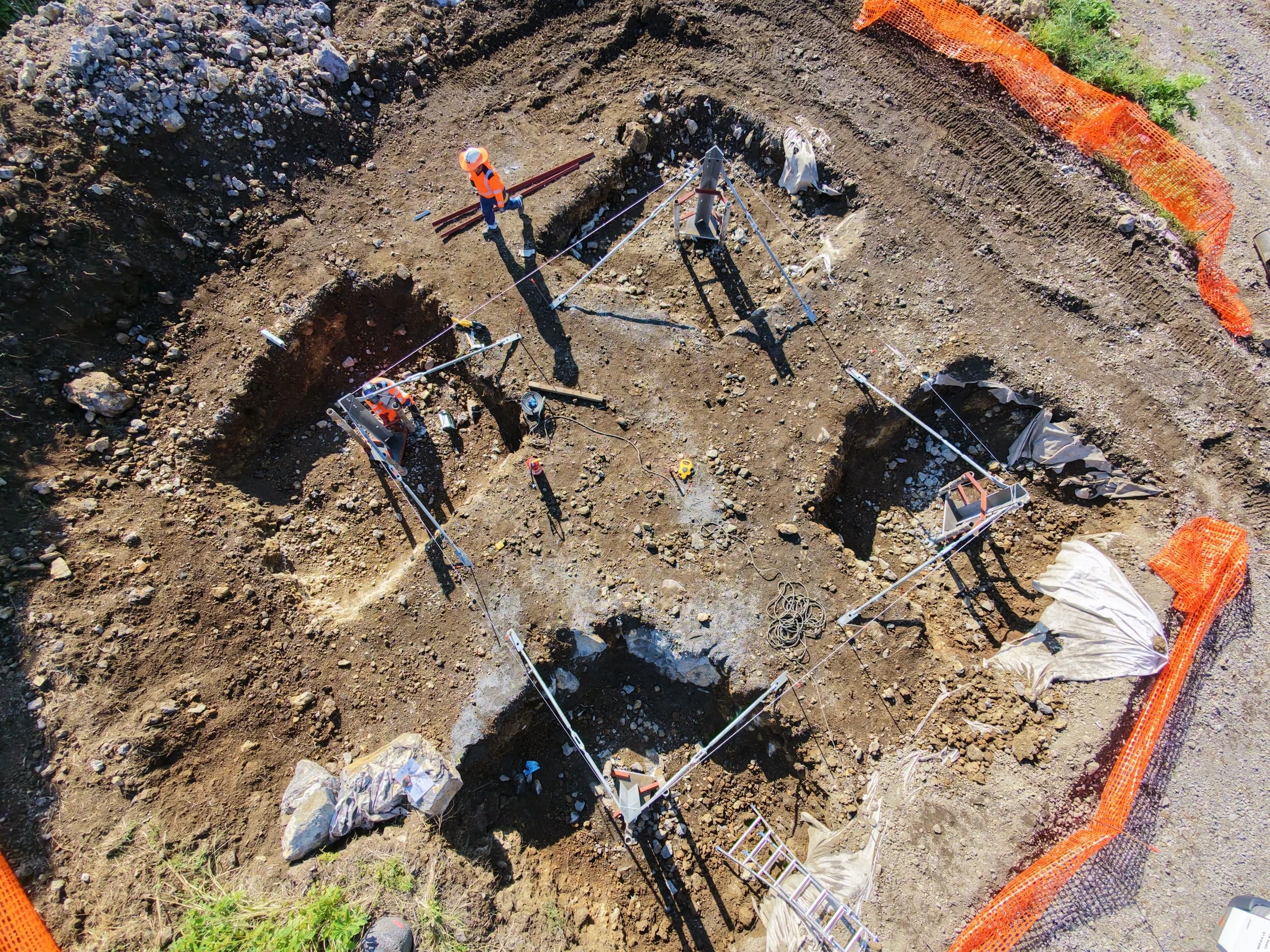 Travaux de fouilles et fondations avec des ouvriers et du matériel dans une zone de sol dénudé, entourée d'une barrière orange.