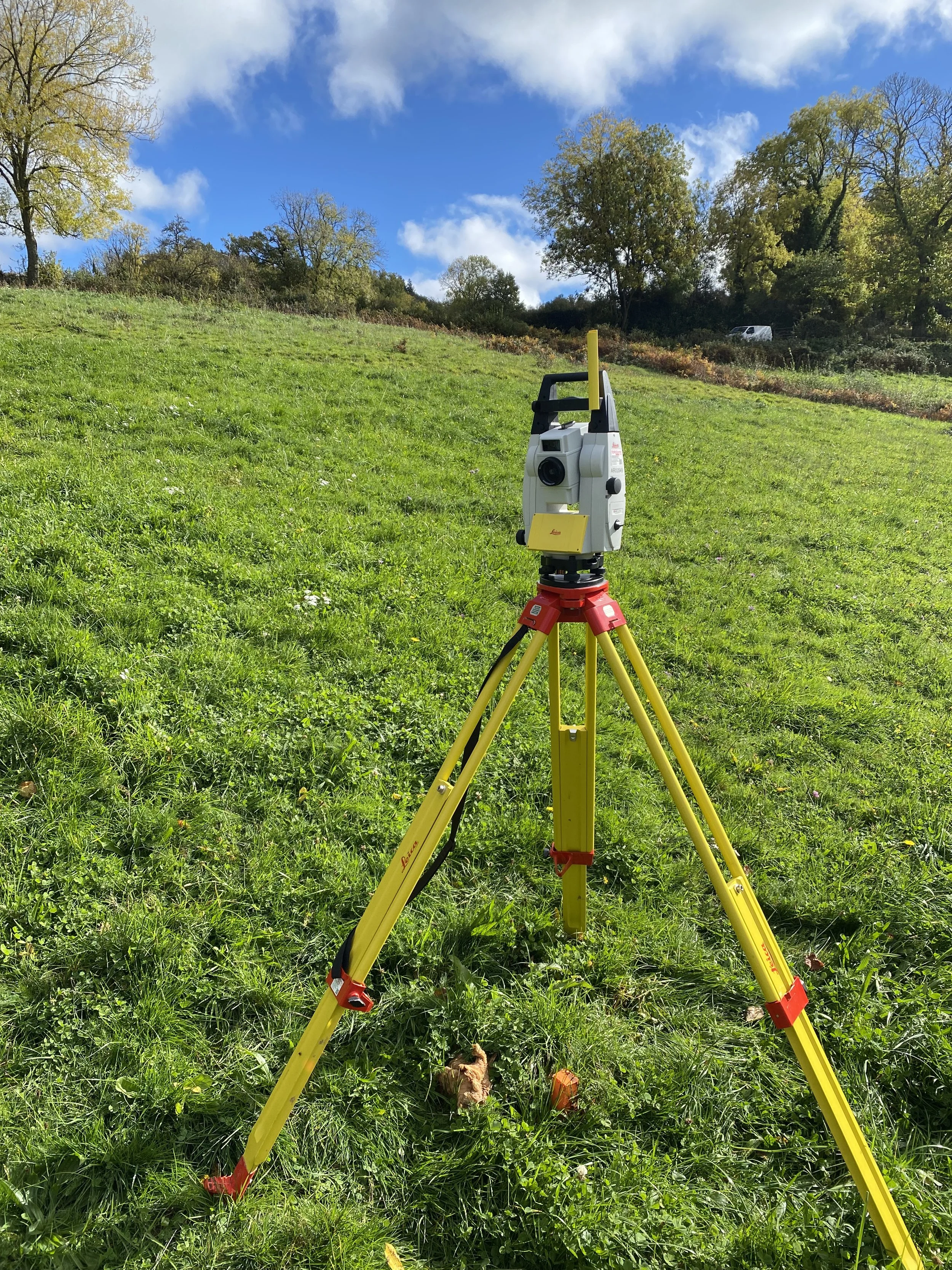 Niveau laser jaune posé sur un sol herbeux en plein air, avec des arbres et un ciel bleu avec des nuages en arrière-plan.