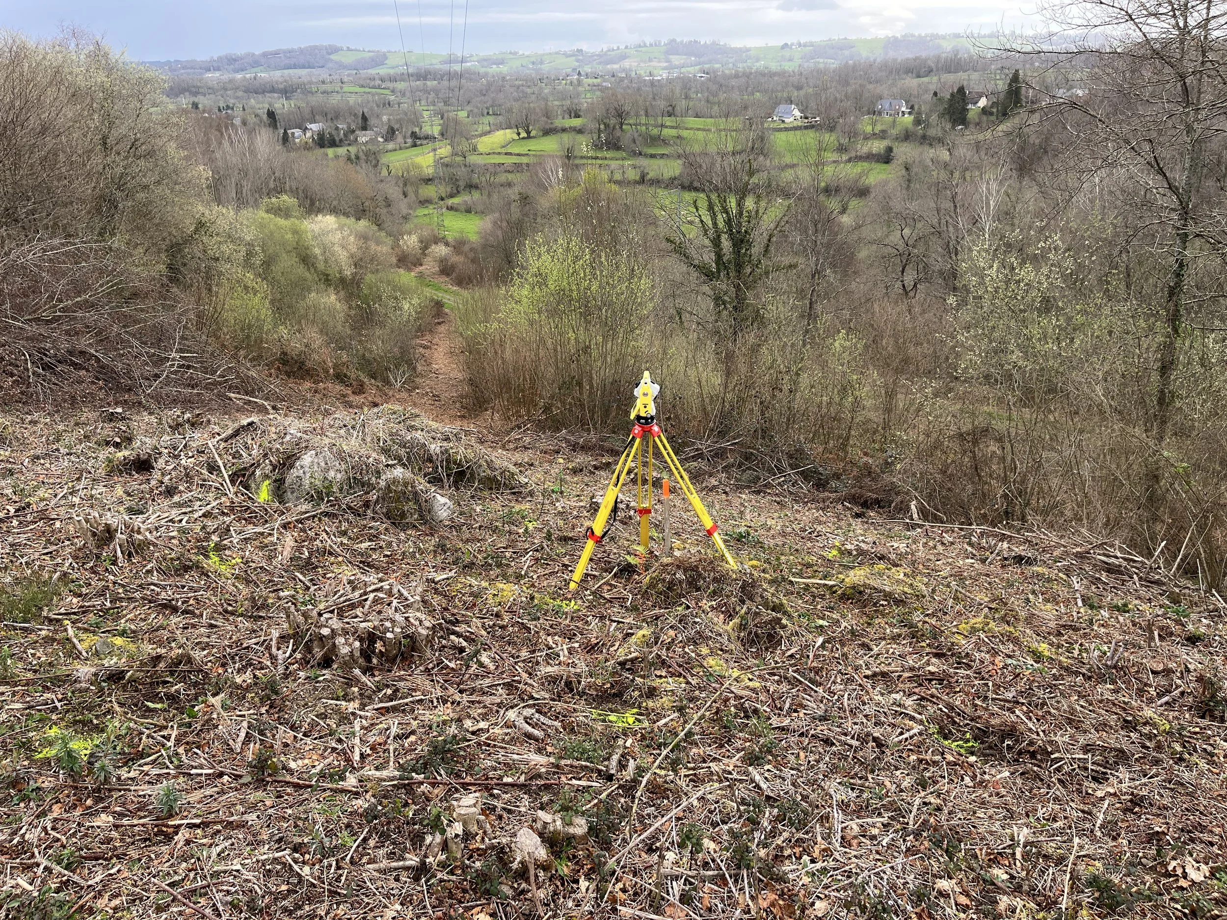 Outil de géomètre avec trépied jaune placé sur un sentier dans une zone boisée, vue sur un paysage vallonné avec des arbres, des prairies et des maisons rurales au loin.
Lebon Services - votre prestataire pour vos chantiers de lignes électriques