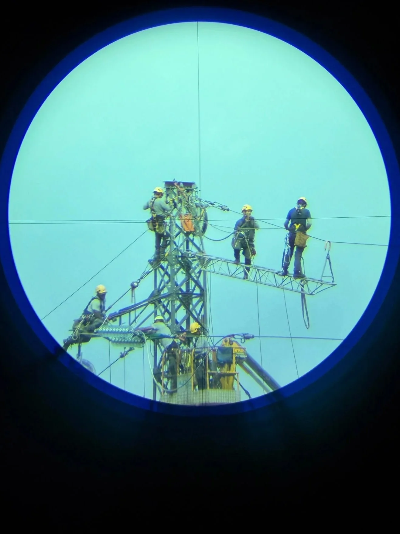 Les travailleurs en uniforme et casque de sécurité effectuent des travaux sur une tour électrique, vus à travers une lunette de visée.