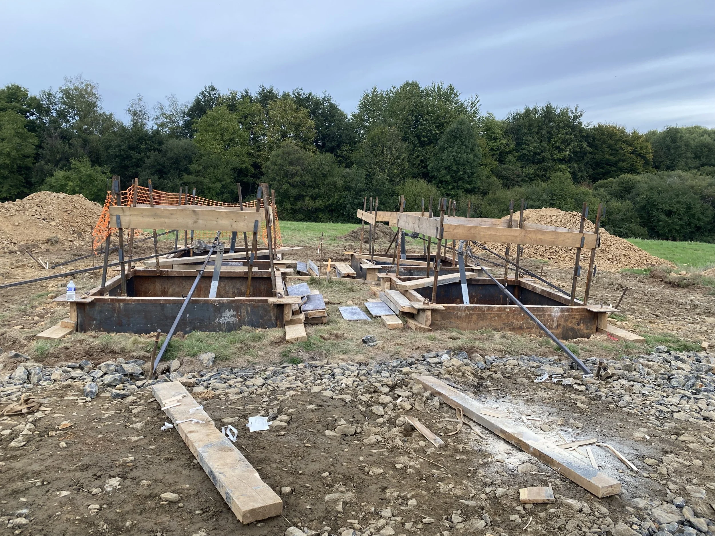 Travaux de construction avec des coffrages en bois pour une fondation, entourés de terre et de roche, avec des arbres en arrière-plan et un ciel nuageux.
Lebon Services