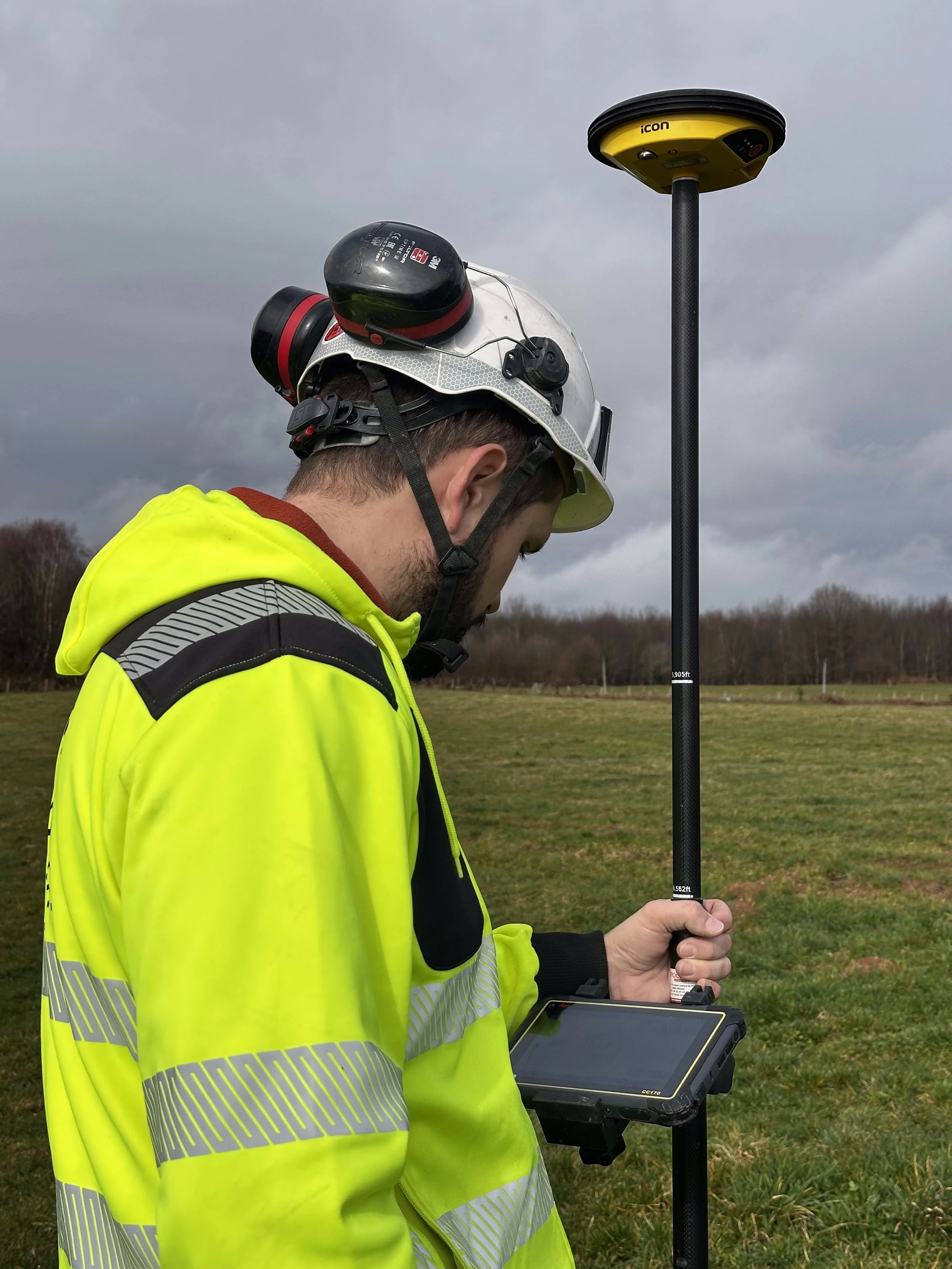 Un homme portant un casque de sécurité et une veste jaune réfléchissante utilise un drone ou un équipement de mesure électronique dans un champ en plein air, avec un ciel nuageux en arrière-plan.