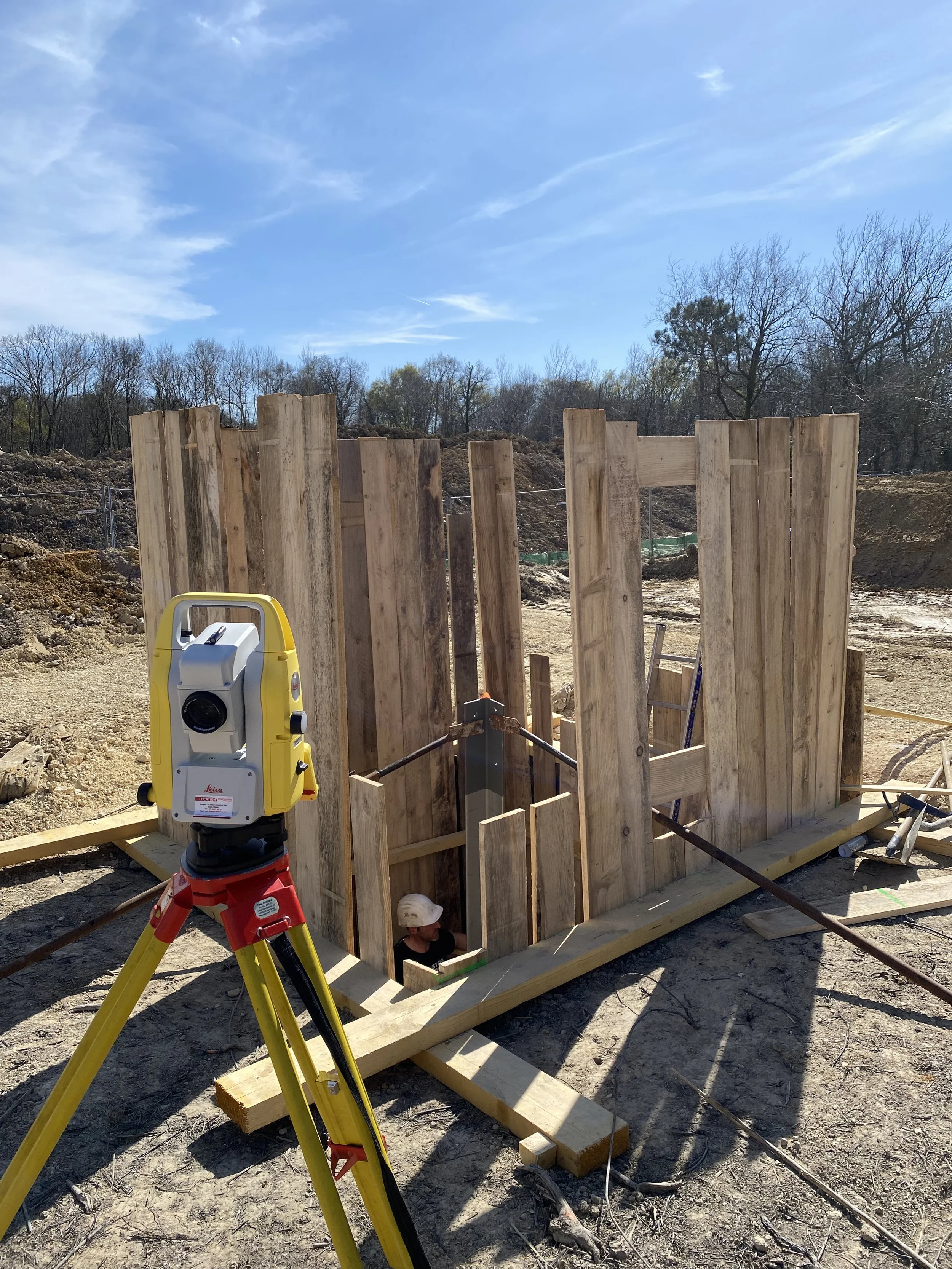 Construction en cours avec des murs en bois, un niveau à laser jaune sur un trépied, et un ouvrier sous le cadre.