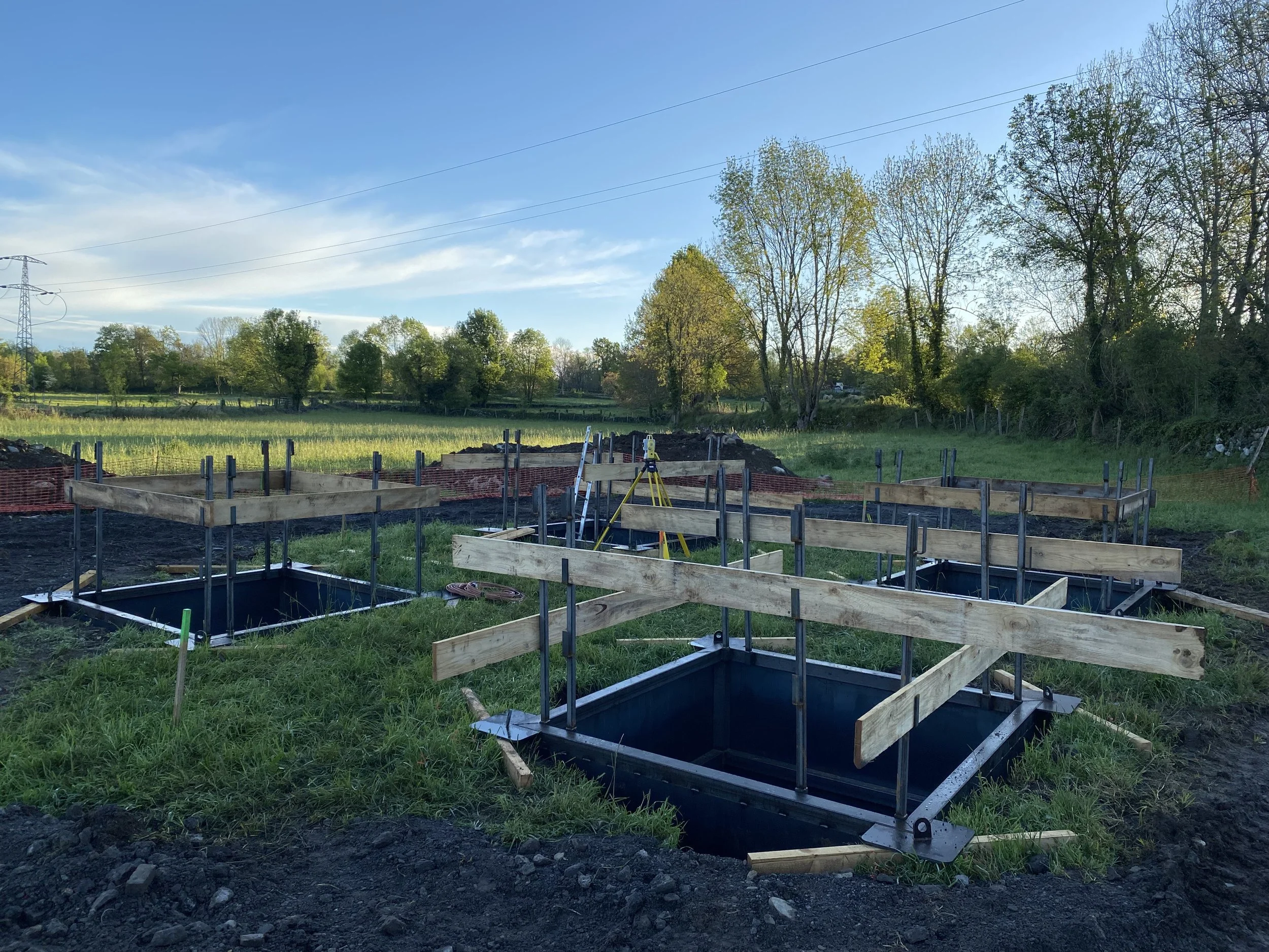 Chantier de construction avec des fondations en cours, des structures en bois et un terrain herbeux entouré d'arbres et un ciel bleu avec quelques nuages.