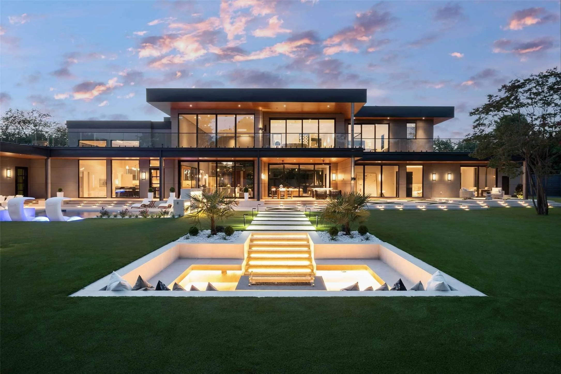 Modern house at dusk with illuminated outdoor yard, stairs leading to a pool, and large glass windows showing interior lighting.