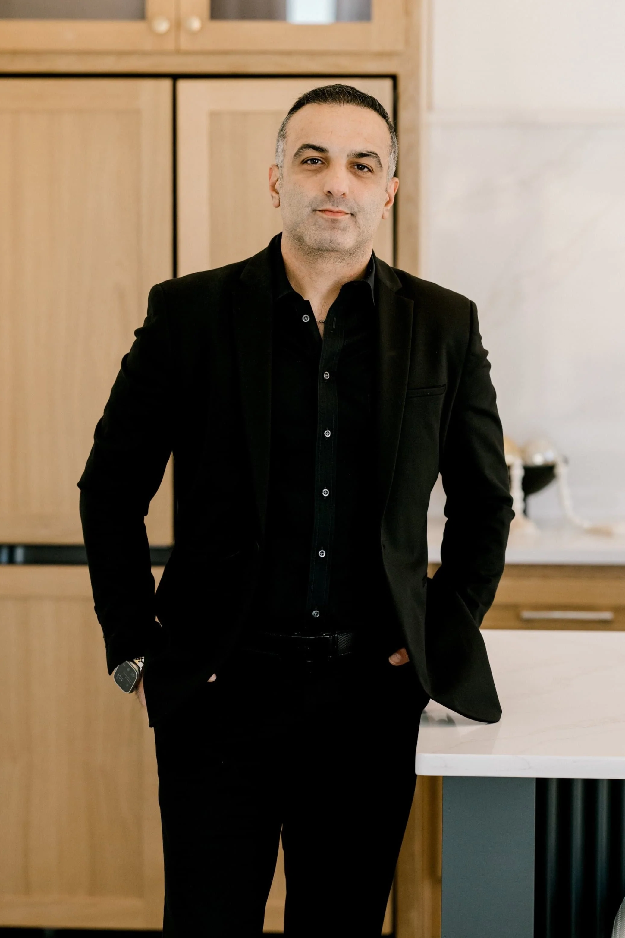 A man wearing a black suit and black shirt standing in a modern kitchen with light wood cabinets and a white marble countertop.