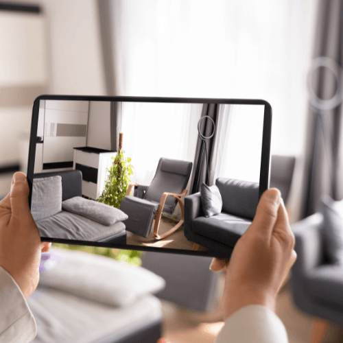 A person holding a tablet while capturing an image or video of a modern living room, which features sleek gray furniture, a wooden rocking chair, and natural light streaming in through sheer curtains.