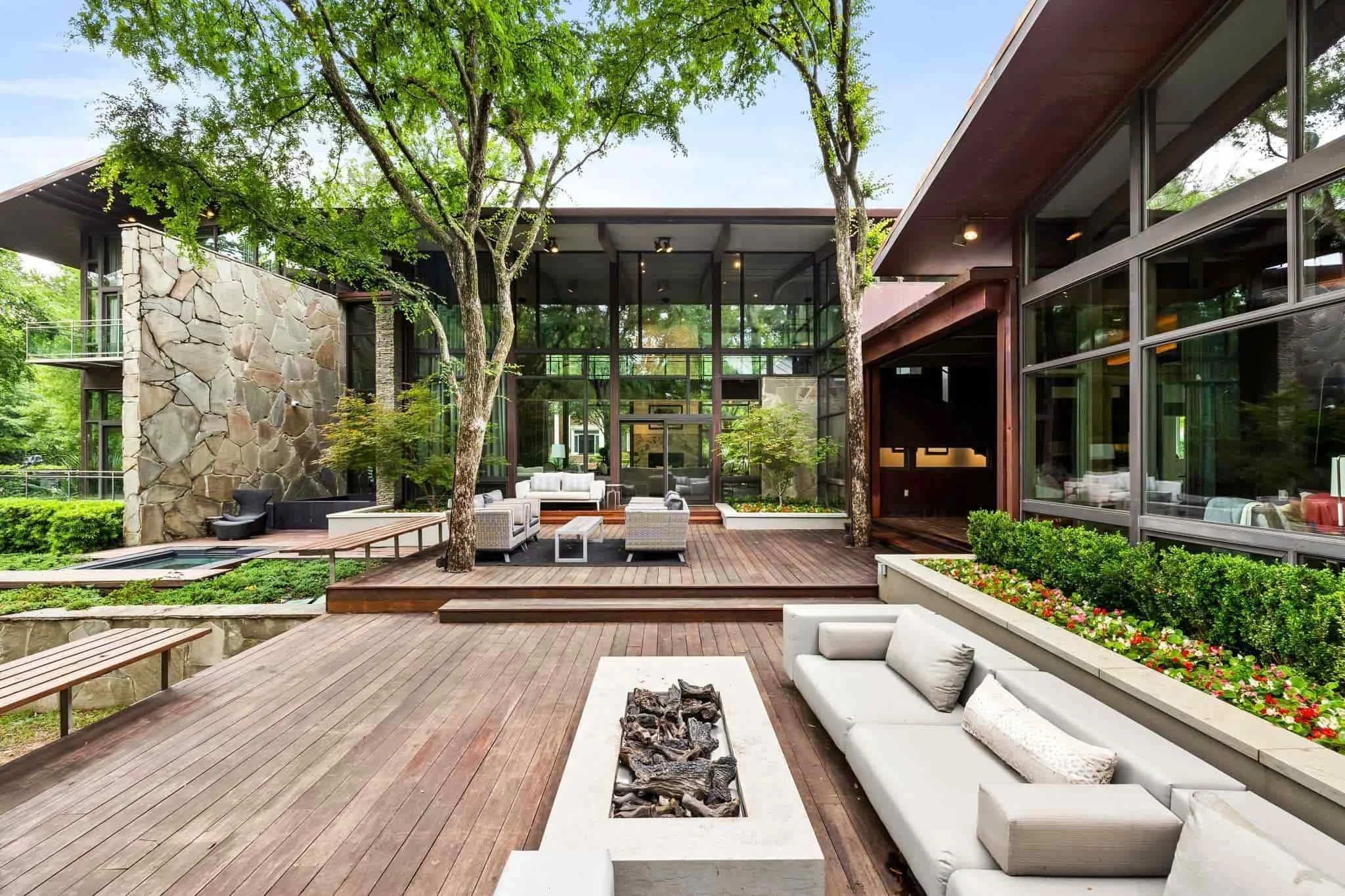 Modern house with large glass windows and wooden deck patio featuring outdoor seating and a fire pit, surrounded by greenery and trees.