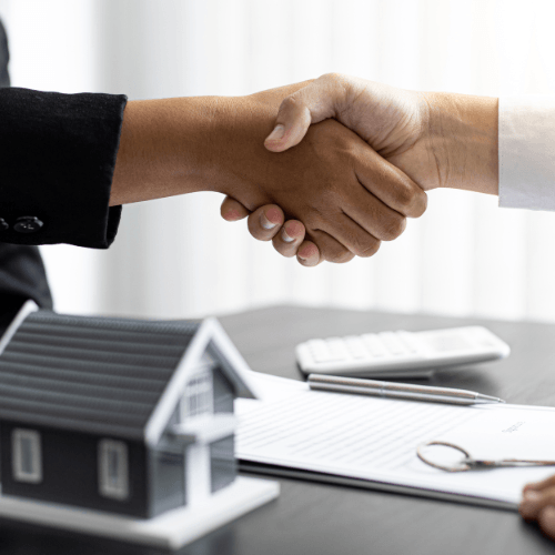 The image shows two individuals shaking hands, symbolizing a successful agreement or deal. In the foreground, there is a small model of a house, representing real estate, and documents on the table