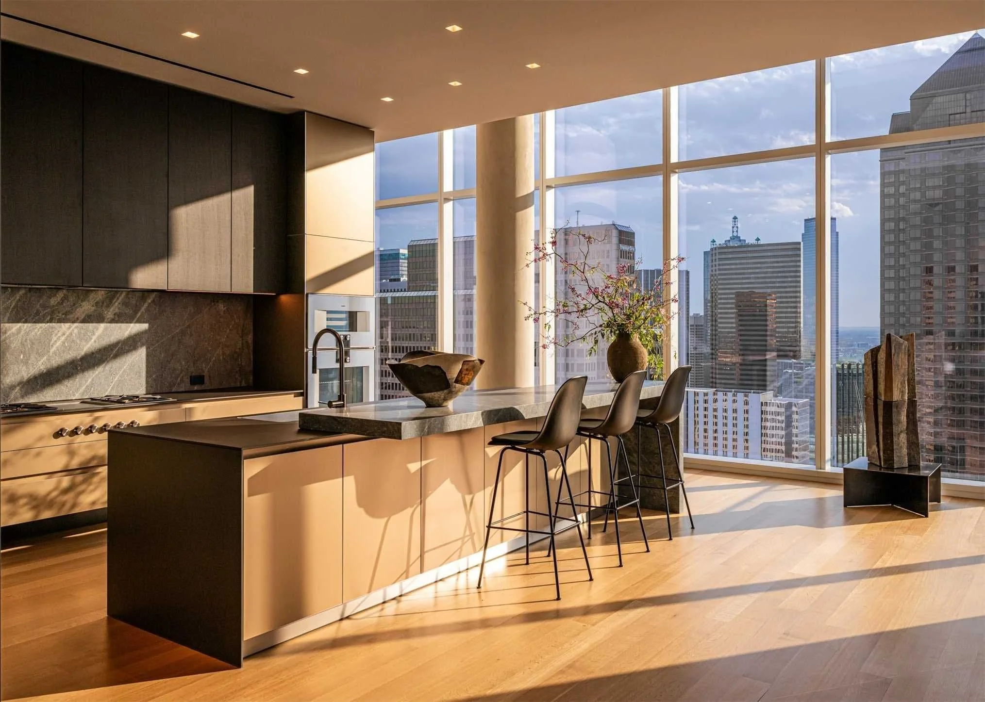 Modern kitchen in high-rise apartment with large windows, city skyline view, kitchen island with three black barstools, decorative bowl and plant on the countertop, floor lamp, and wooden floors.