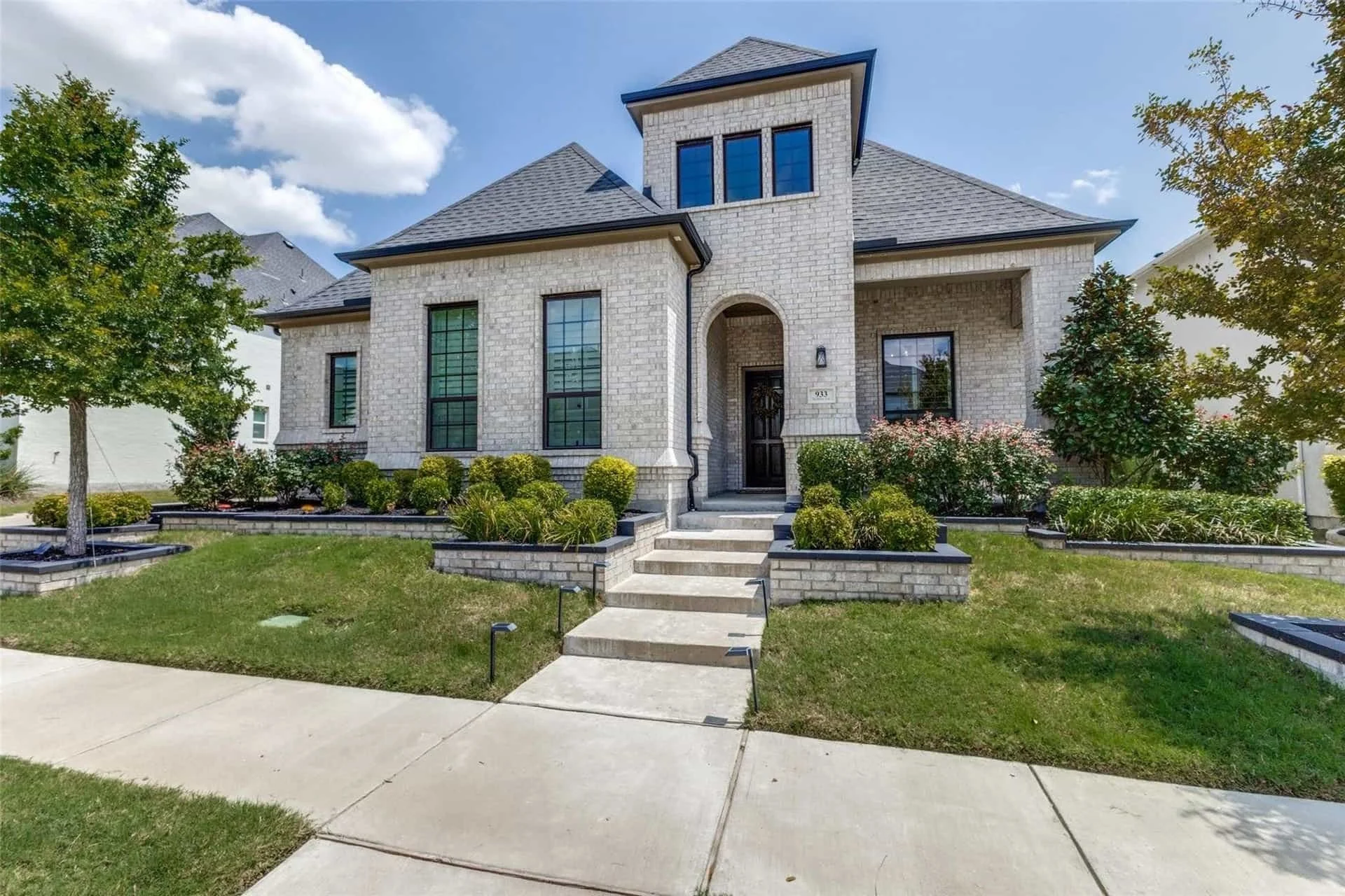 A modern two-story house with a brick exterior, a dark front door, and large windows, surrounded by a manicured lawn and landscaped garden with bushes and small trees under a blue sky with some clouds.
