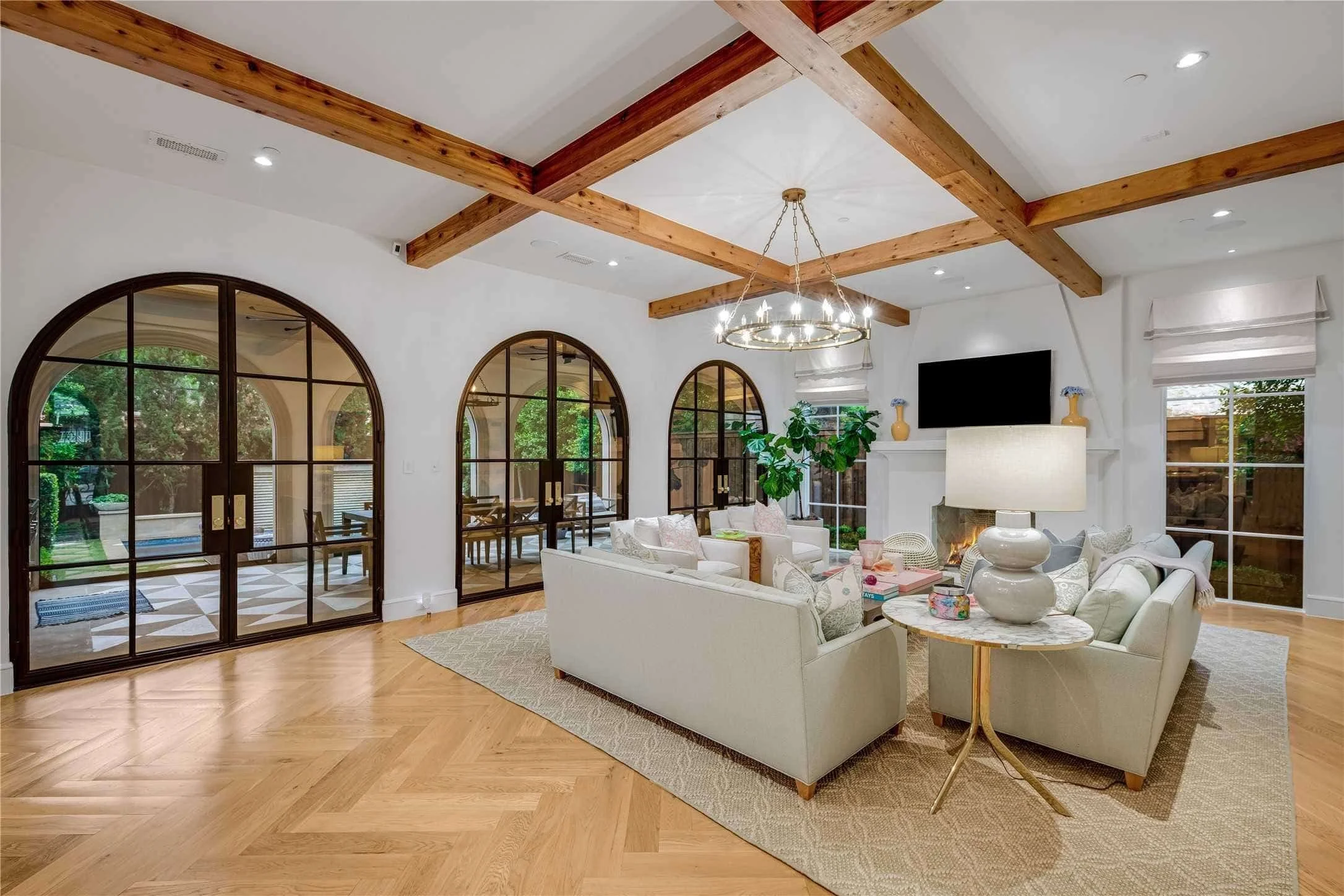 Elegant living room with white walls, wooden beams on the ceiling, and a large area rug. There are white sofas, a side table with a lamp, a fireplace, a flat-screen TV, and large arched windows showing an outdoor patio.
