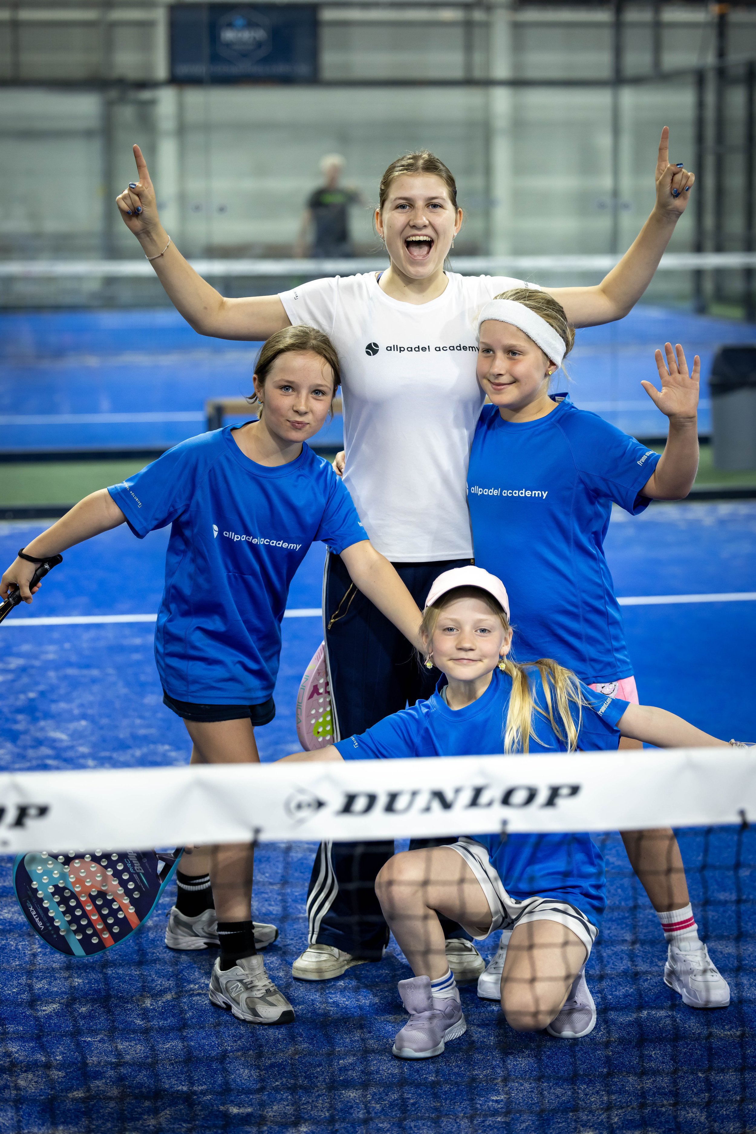 Groep kinderen en een vrouw op een padelbaan, allemaal in sportkleding. Kinderen dragen blauwe shirts met logo 'allpadel academy' en de vrouw draagt een wit shirt met hetzelfde logo. Ze lijken blij en poseren voor een foto.