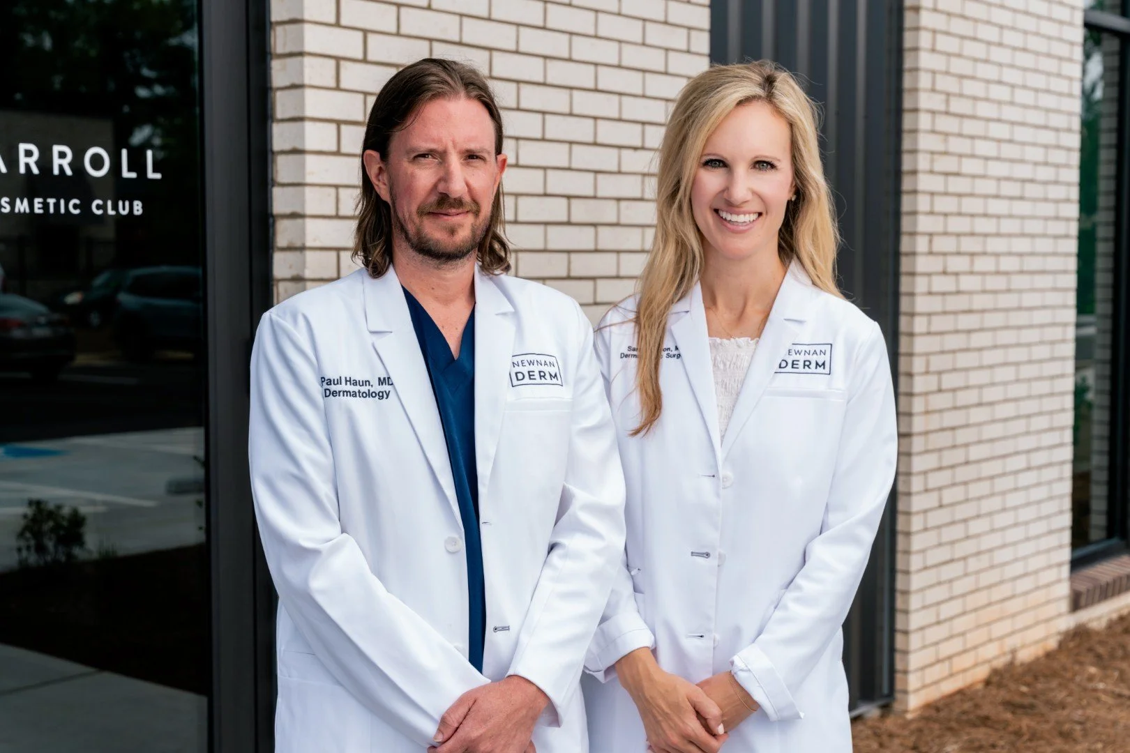 Two dermatologists in white coats standing outside a building with a brick wall and glass window. One man with long hair and a beard, and one woman with long blonde hair, smiling.