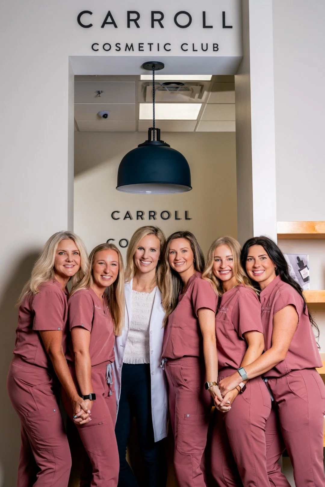 Group of six women, five in mauve medical scrubs and one in a white coat, standing together inside a cosmetic clinic with a sign reading 'Carroll Cosmetic Club' above and behind them, smiling at the camera.
