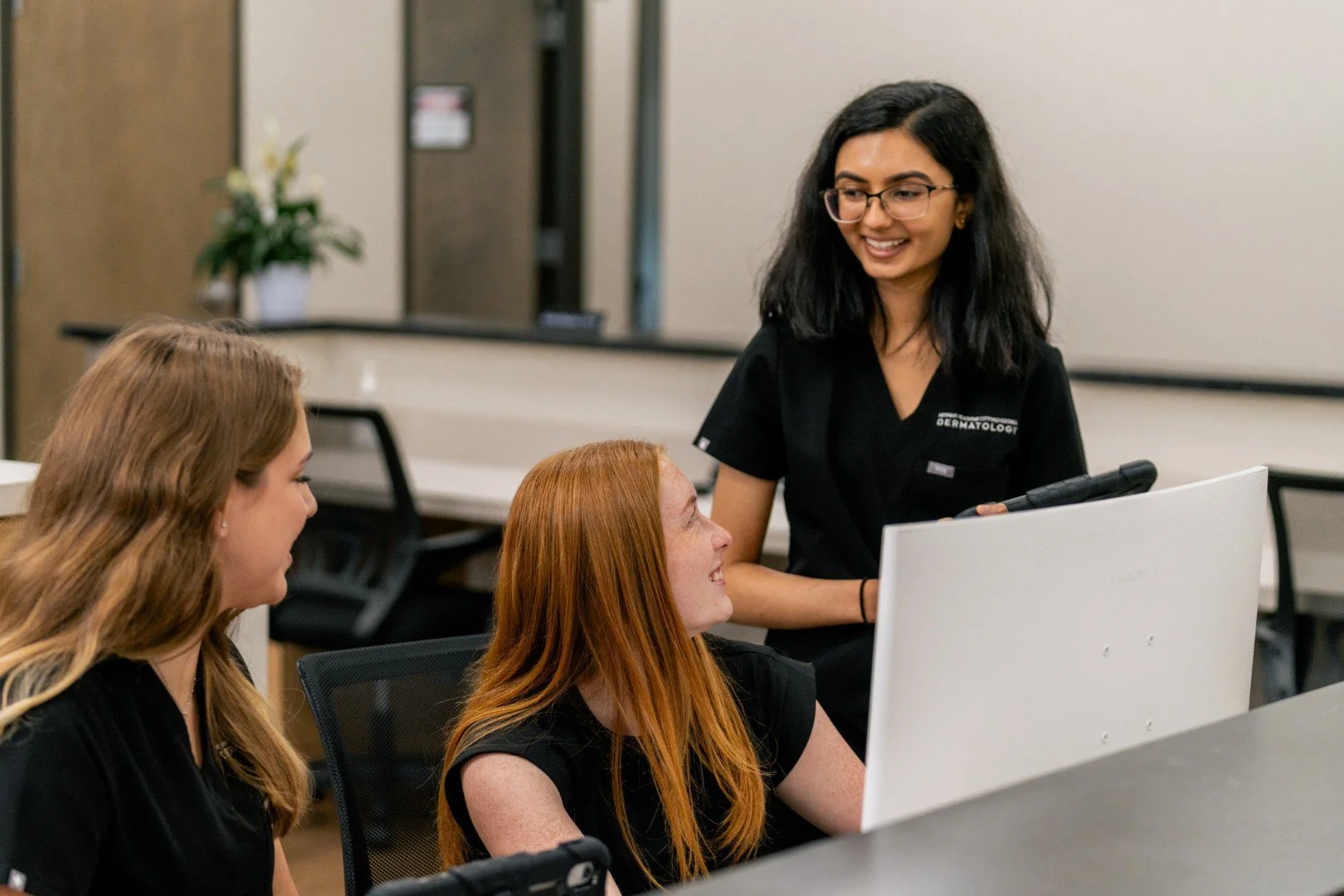 A nurse or healthcare professional in black scrubs with glasses smiling while talking to two young women sitting at a desk in an office or classroom setting.