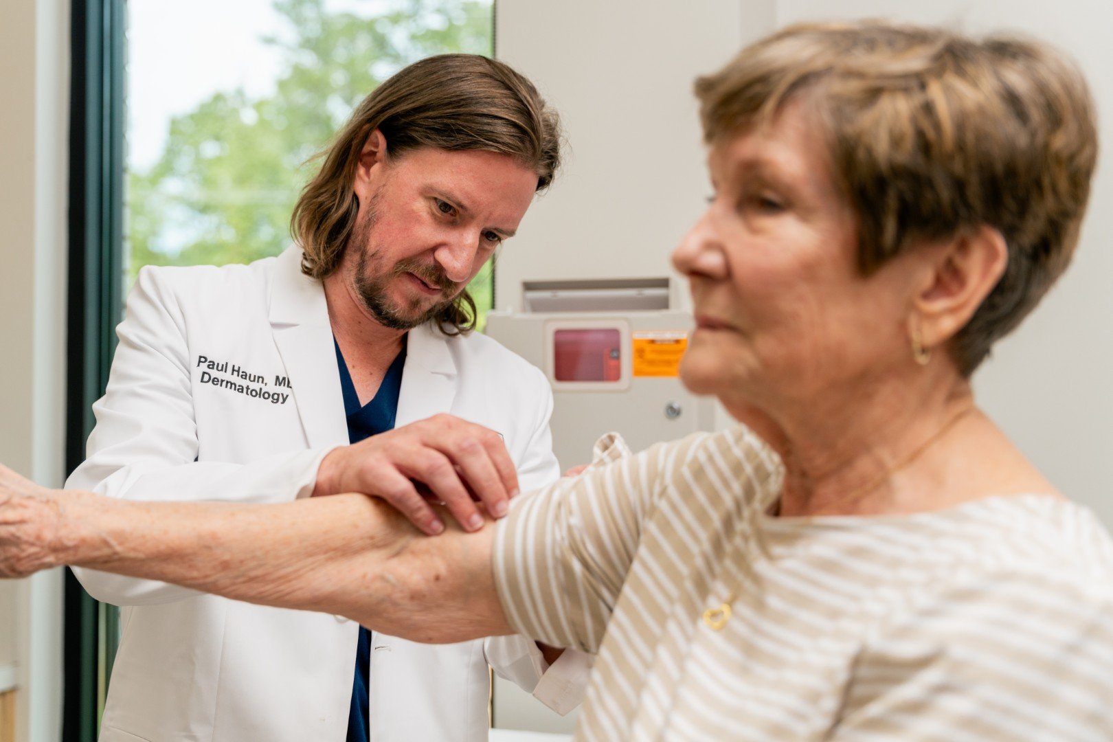 A dermatologist in a white coat examines an elderly woman's arm in a medical office.