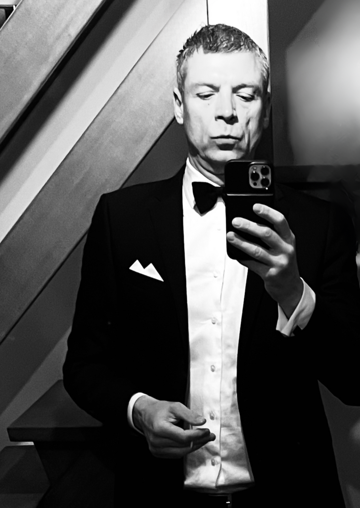 Man in een zwart pak en een zwarte strik, poserend voor een spiegel in zwart-wit. Hij neemt een foto met een smartphone.