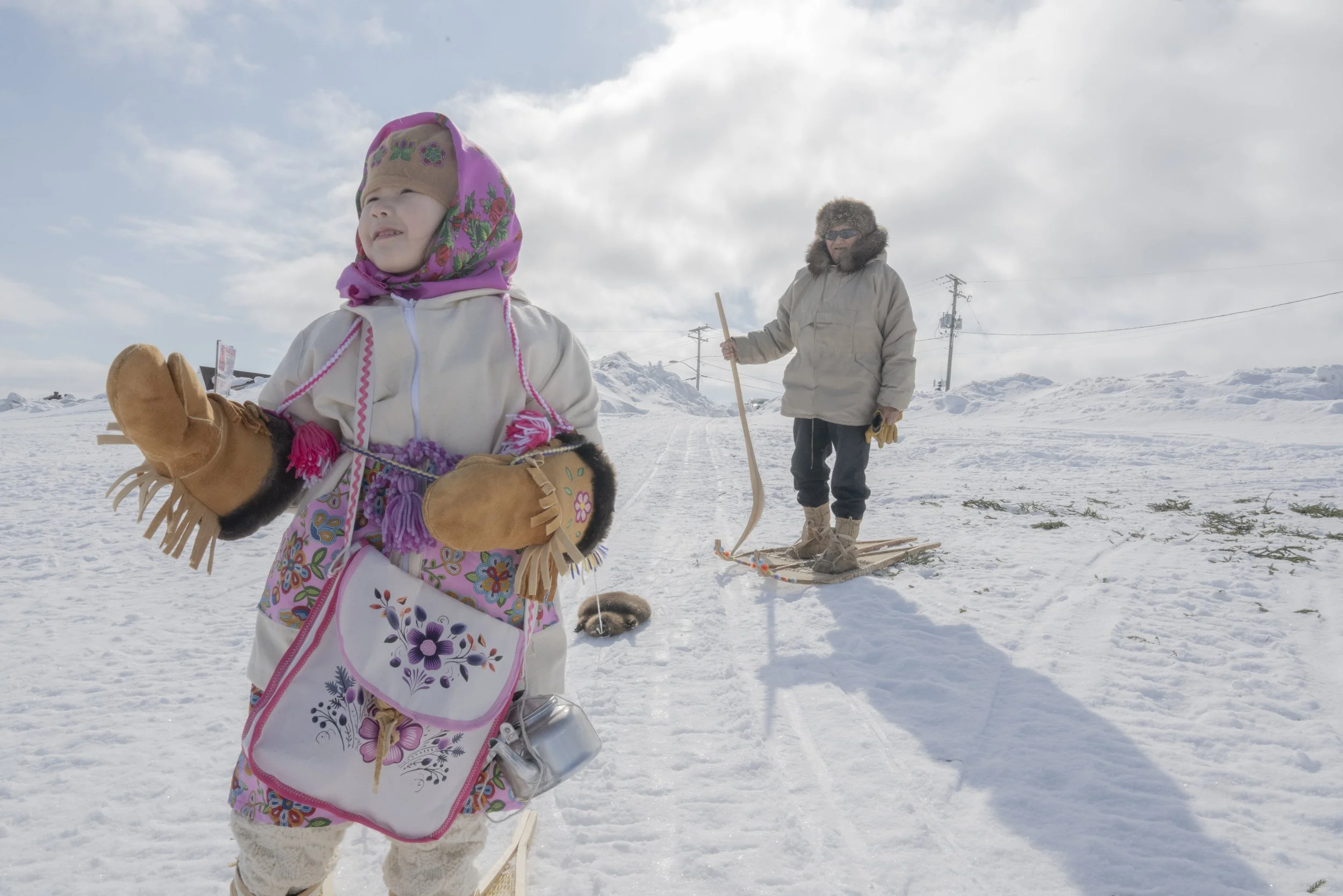 A young girl dressed in traditional Cree clothing, with a local Elder standing behind her.