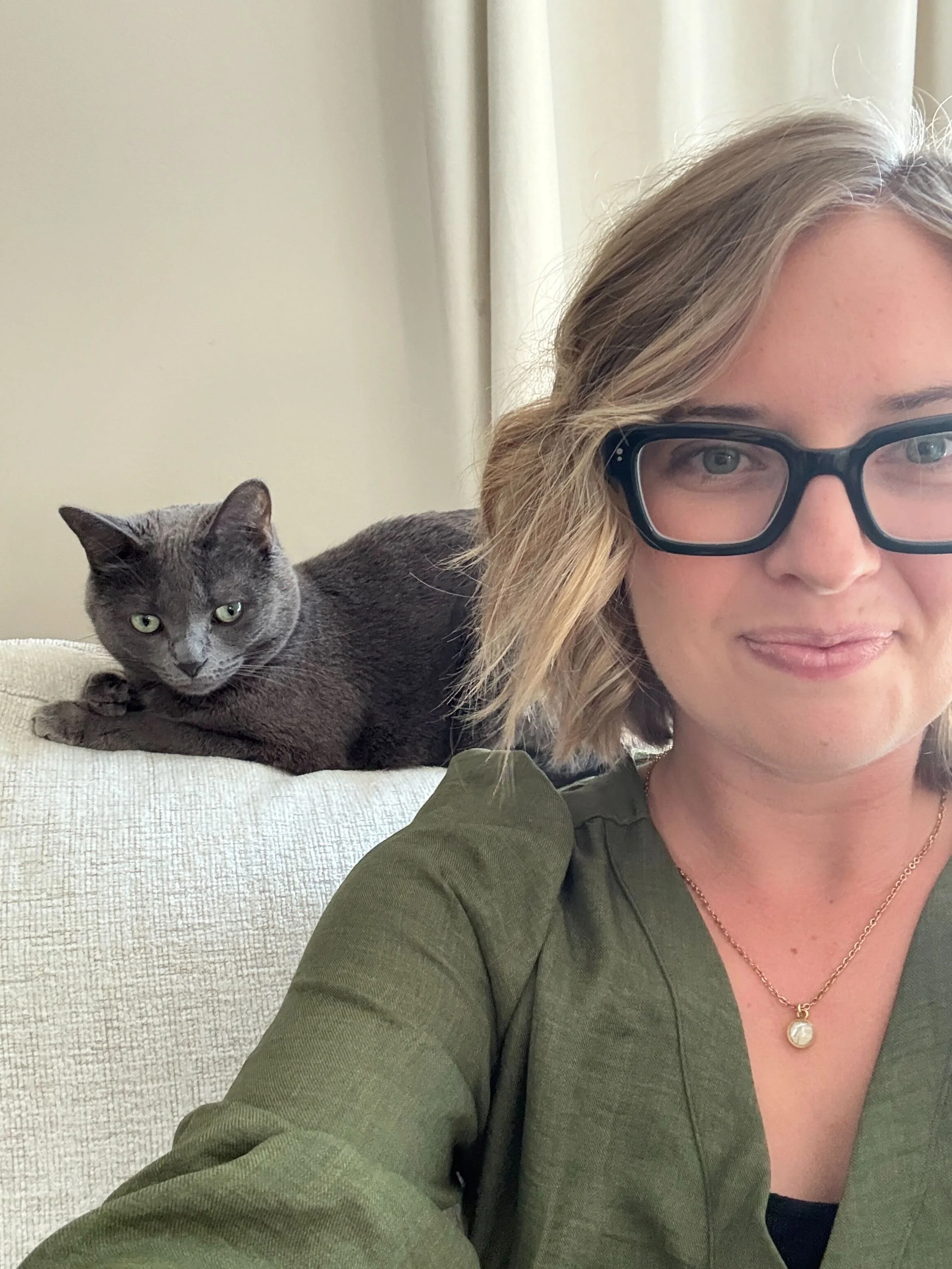 A woman with light skin, blonde wavy hair, and black glasses taking a selfie. Next to her, a gray tabby cat with green eyes lying on a light-colored fabric surface, staring directly at the camera. A cream-colored curtain is visible in the background.