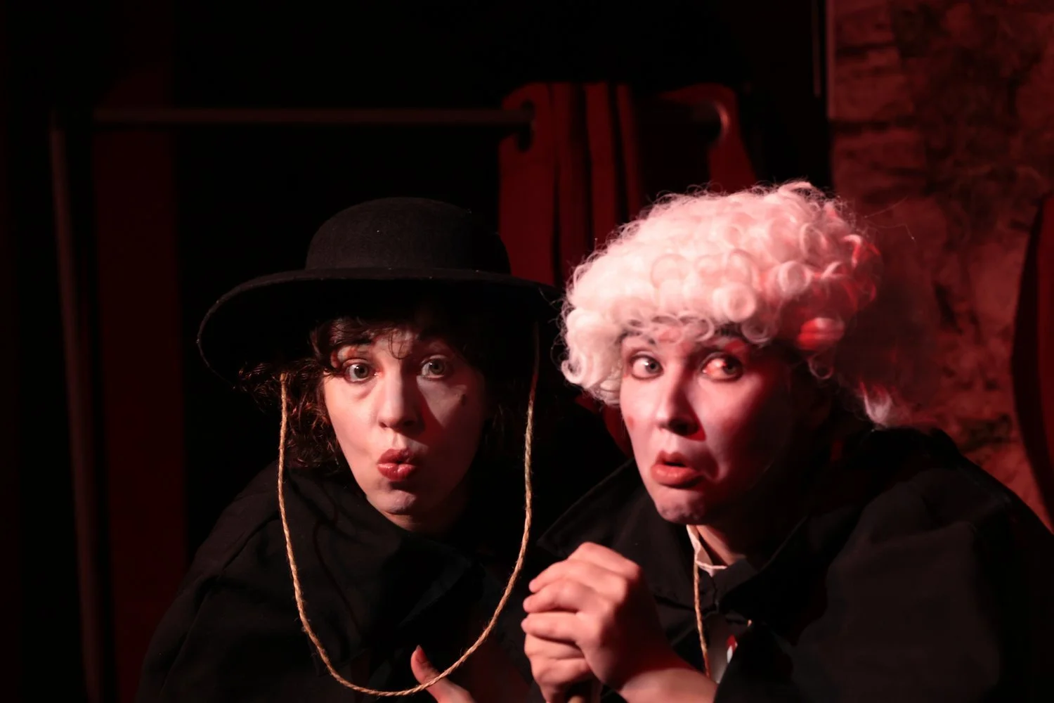 Deux artistes en costume de théâtre, un homme avec perruque blanche bouclée et une femme avec un chapeau noir, respectant la scène dans une ambiance dramatique.