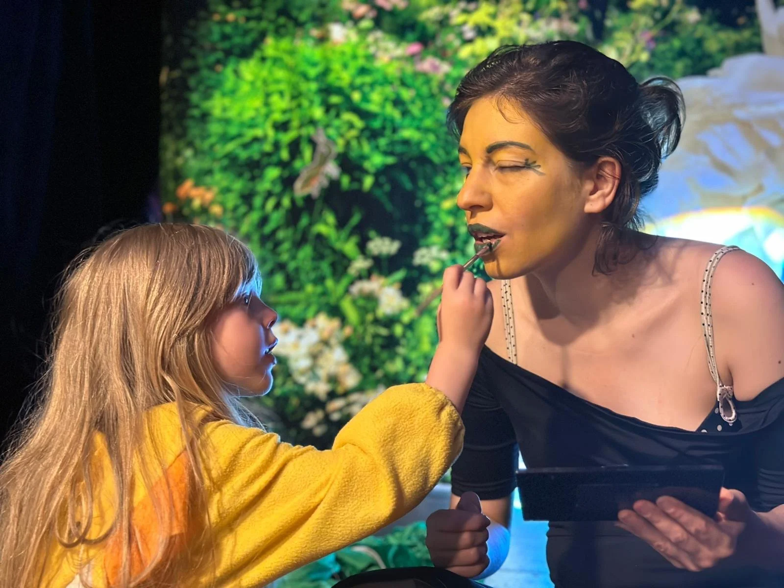 Une jeune fille maquille une femme avec un pinceau. La femme a du maquillage artistique autour de l'œil. Le fond montre une image colorée de verdure et de fleurs.