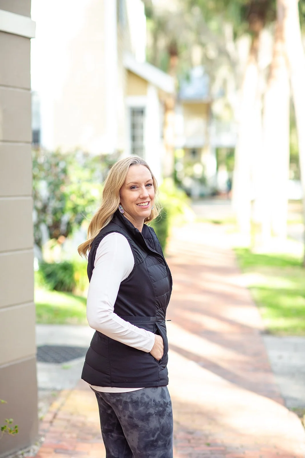 A woman with blonde hair smiling outdoors on a sidewalk, wearing a white long-sleeve shirt, black vest, and dark patterned pants.