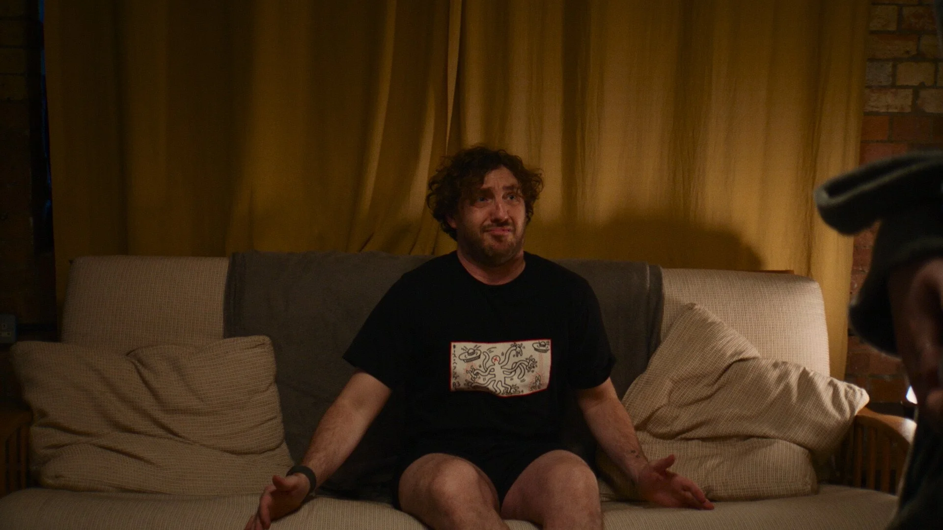 Seann Walsh on a beige couch with beige cushions in a dimly lit room, wearing a black T-shirt with a white and red design, and expressing a distressed or confused emotion.