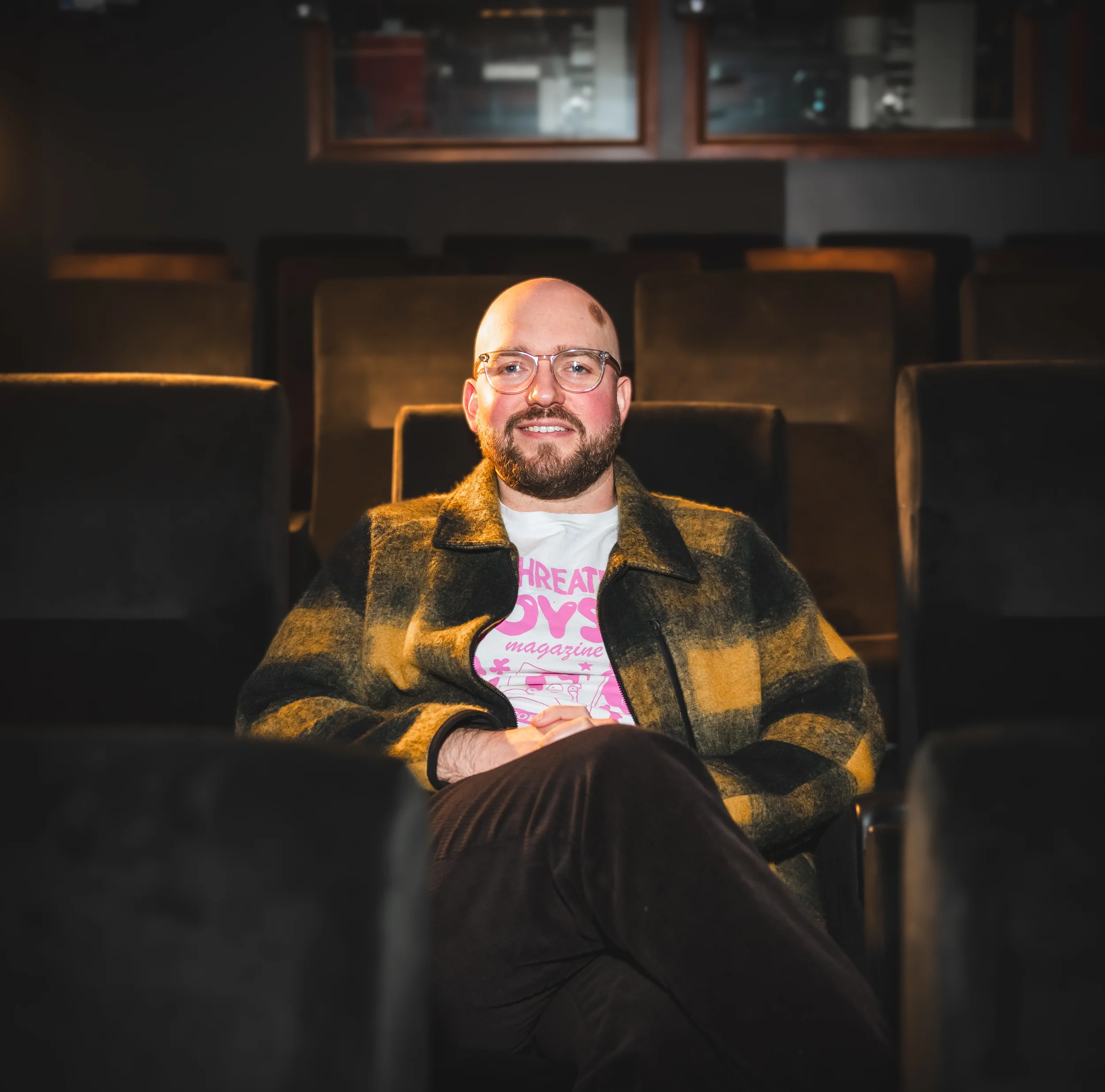 A man with glasses and a beard, wearing a plaid jacket and a graphic t-shirt, seated in a dimly lit theater or auditorium with empty seats around him.