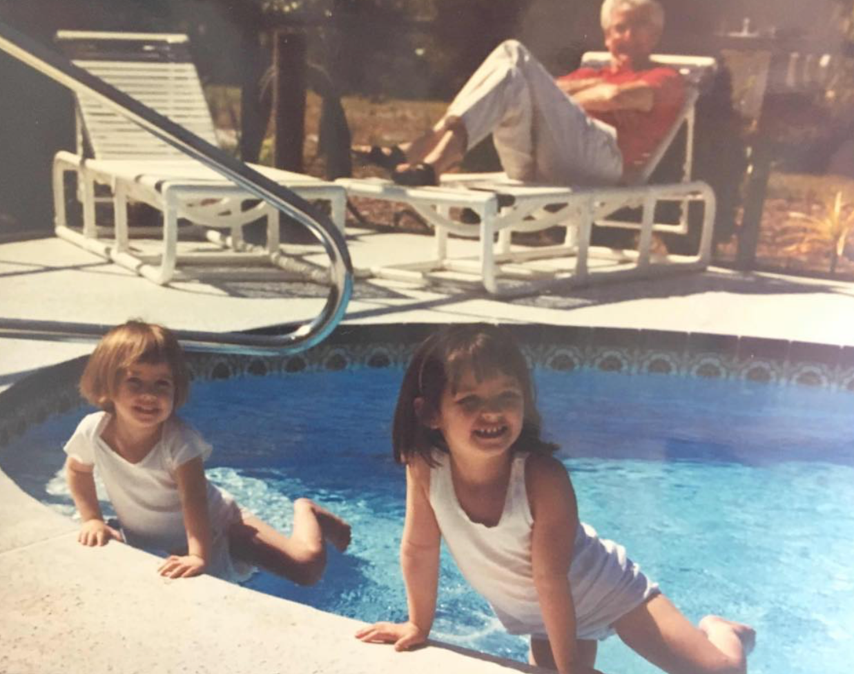 Two young girls in white tank tops and shorts sitting in a backyard swimming pool, smiling at the camera. An elderly woman is relaxing on a lounge chair, sitting with her legs stretched out and crossed, with a dog lying on her lap. The setting appears to be a sunny day in a backyard.