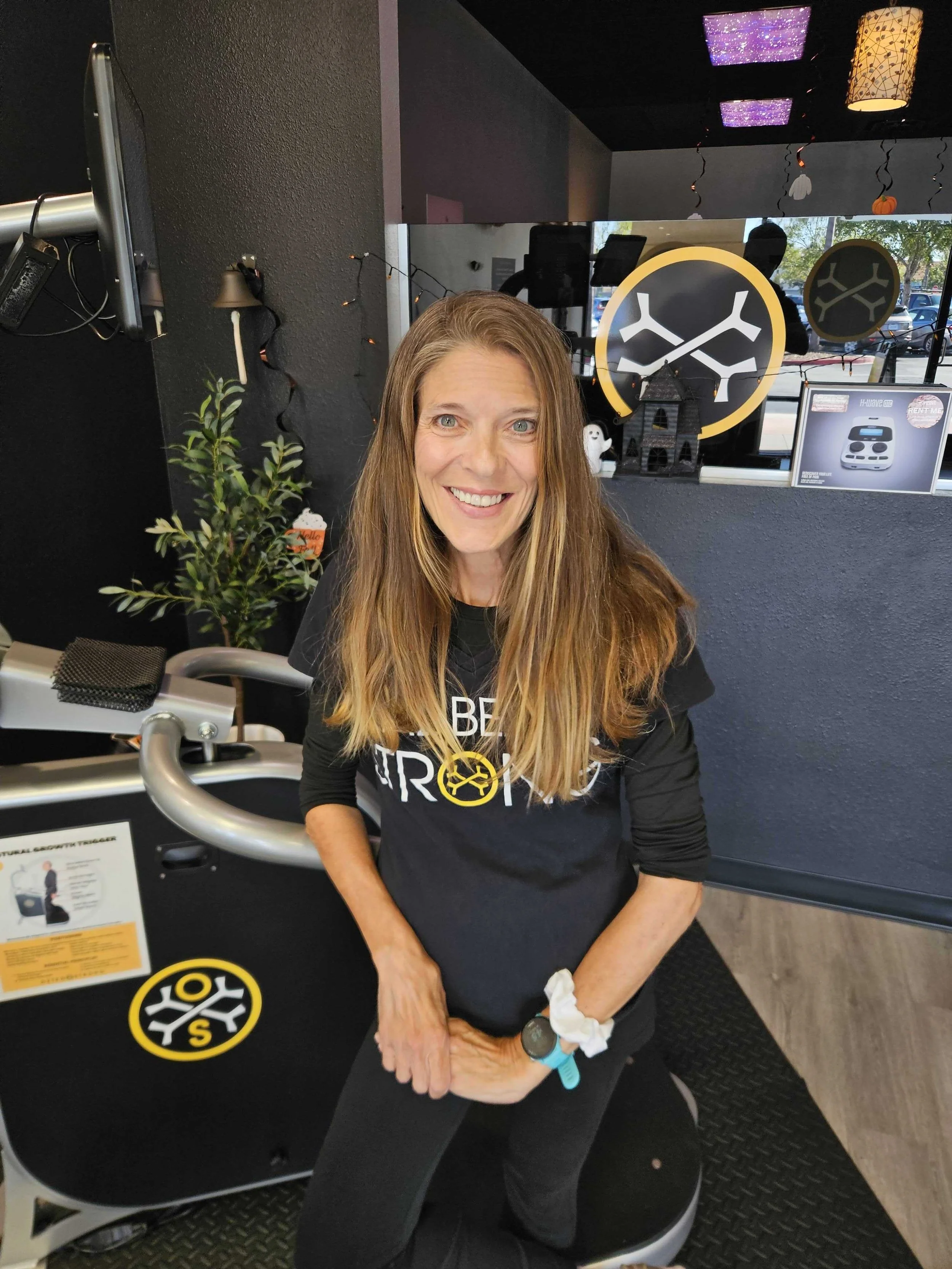 A woman smiling in front of a backdrop decorated with black and gold balloons and confetti. She is wearing glasses and a black T-shirt that reads 'SEAL BEACH STRONG' with a yellow emblem.