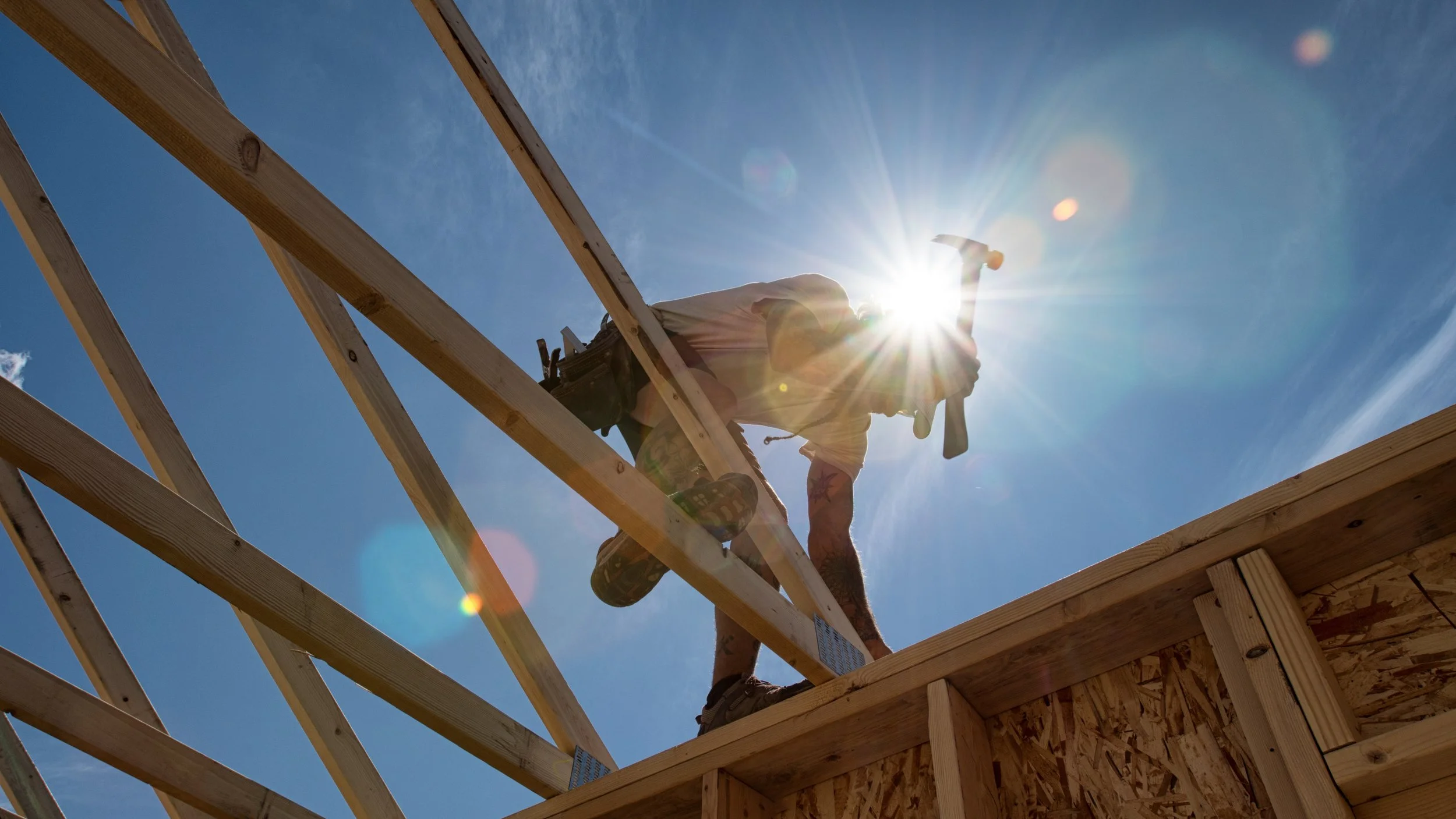 A construction worker installing wooden roof rafters on a building under a bright, sunny sky