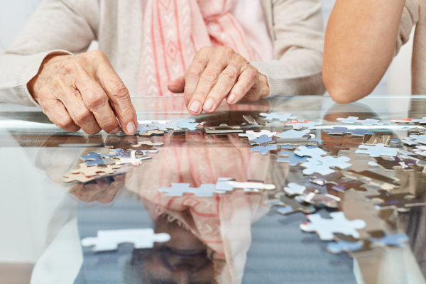 An older woman working on a puzzle with someone beside them helping.