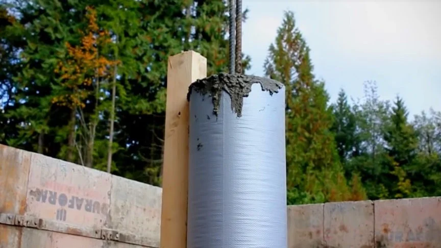 Close-up of a cylindrical metal pipe with wet concrete on top, mounted on a wooden post at a construction site with trees in the background.