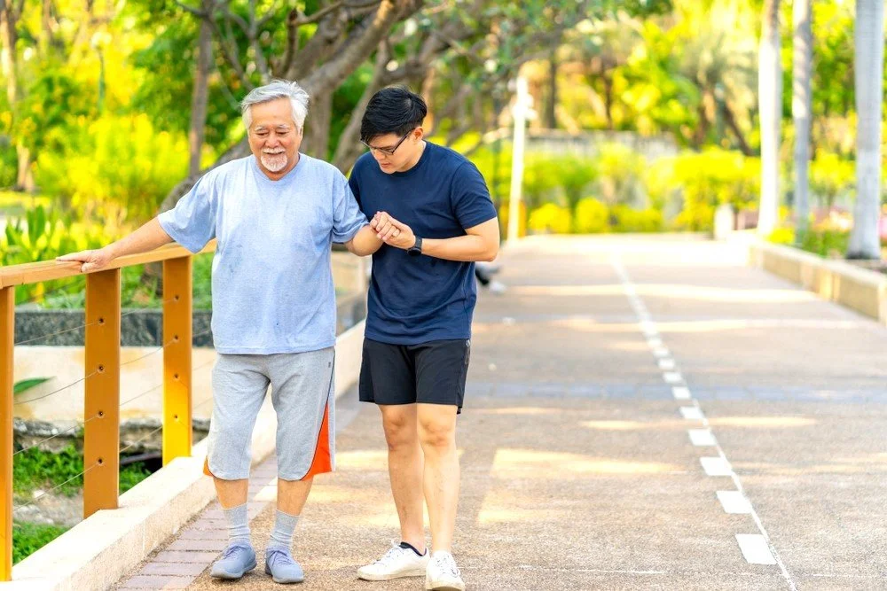 A young son helps his father who is living with dementia by taking a walk along a park path under trees on a sunny day.