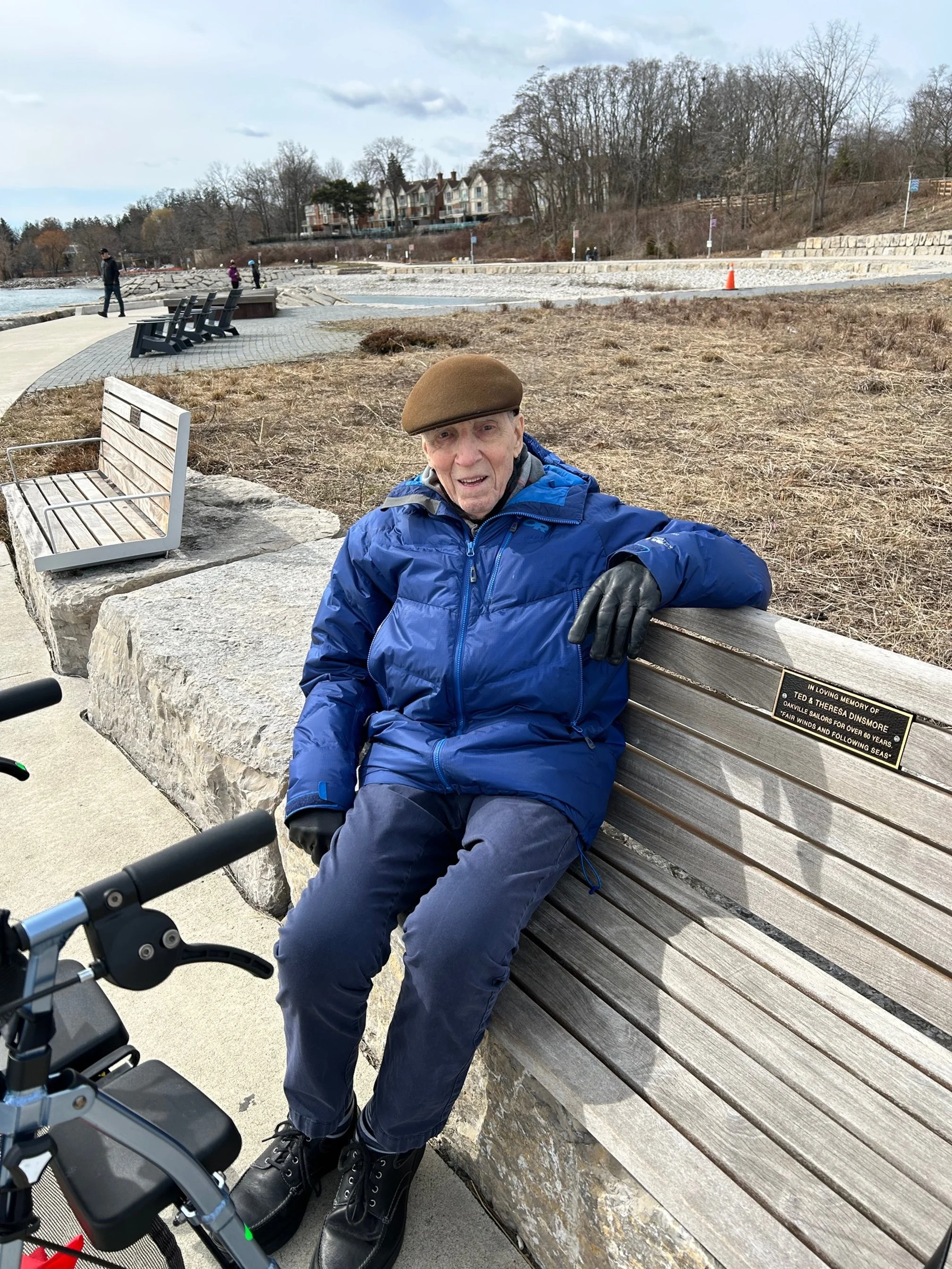An elderly man who is living with dementia is sitting on a bench near the water, wearing a brown cap, blue jacket, black gloves, and black shoes, with a mobility scooter in front of him, in a park or waterfront area.