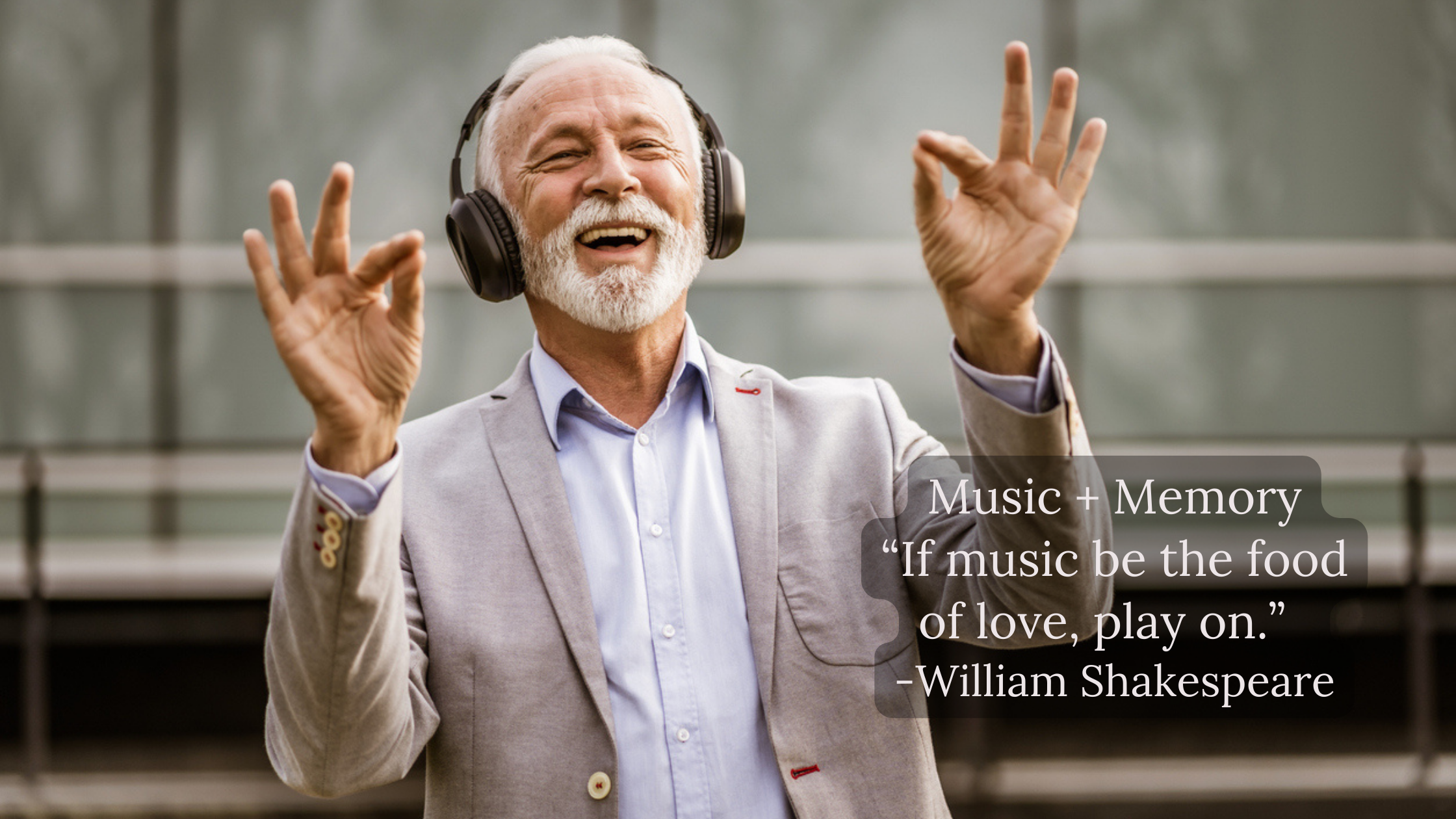 An elderly man with white hair and a beard, wearing a light gray blazer and a blue shirt, is smiling and enjoying music with large black headphones. He is making a gesture with both hands, forming the 'OK' sign, and appears to be very happy. The back