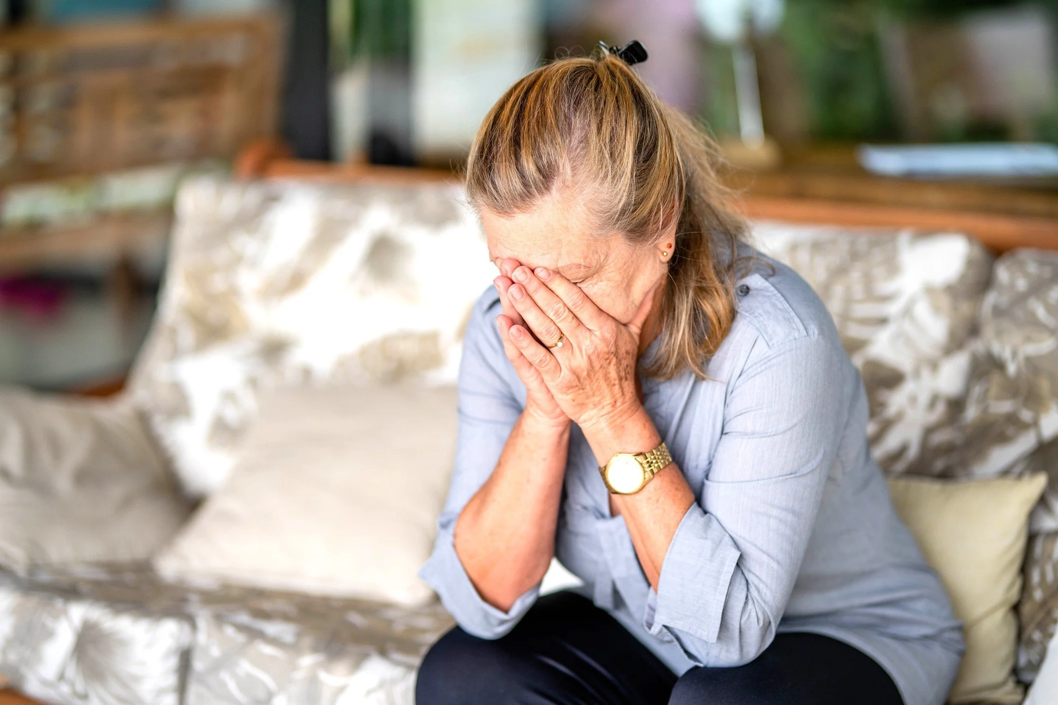 An elderly woman sitting on a sofa covering her face with hands, distressed or emotional who is a primary caregiver for her husband