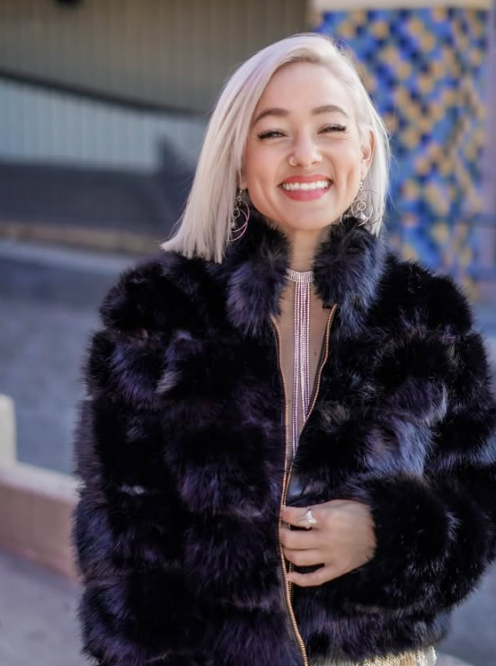 A young woman with platinum blonde hair smiling outdoors while wearing a purple faux fur jacket and large earrings.