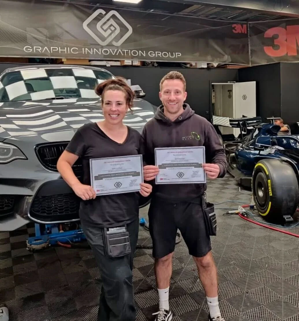 Two people holding certificates in a garage with cars, including a racing car, behind them. There is a large flag or banner above them with the logo and text 'GRAPHIC INNOVATION GROUP' and the 3M logo visible.